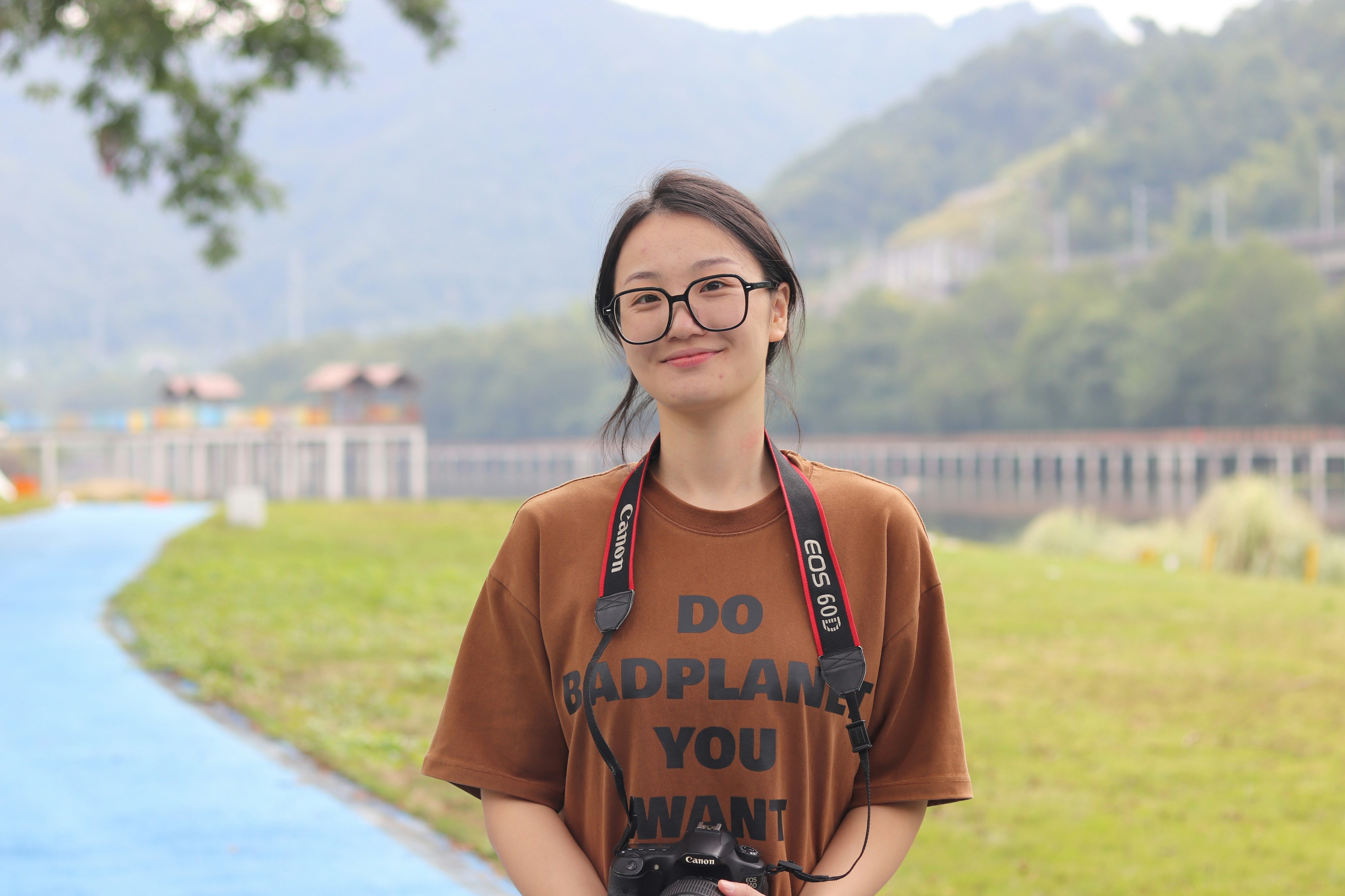 A young woman with a camera stands outdoors near mountains.