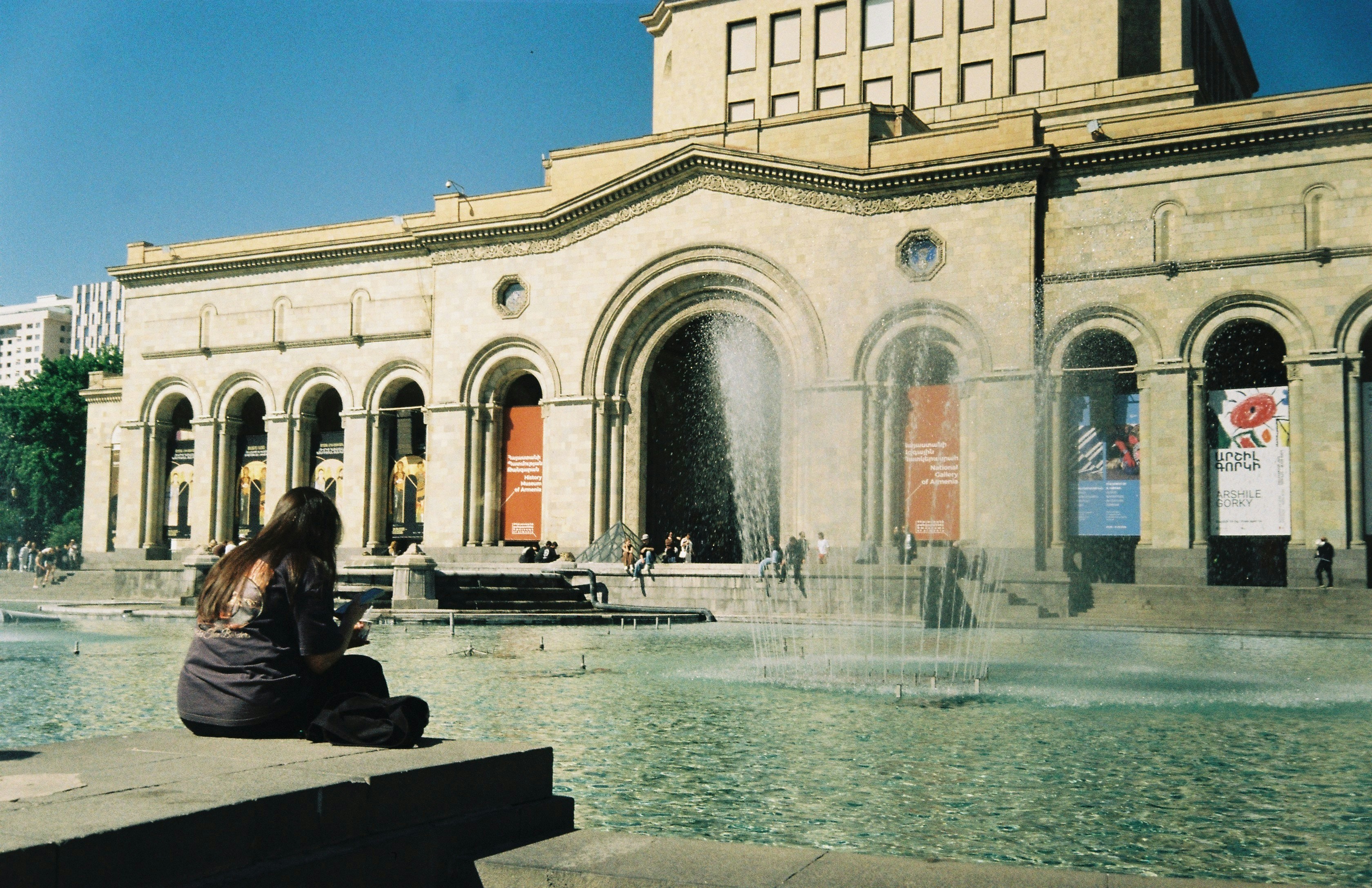 A person sits by a fountain and a grand building.