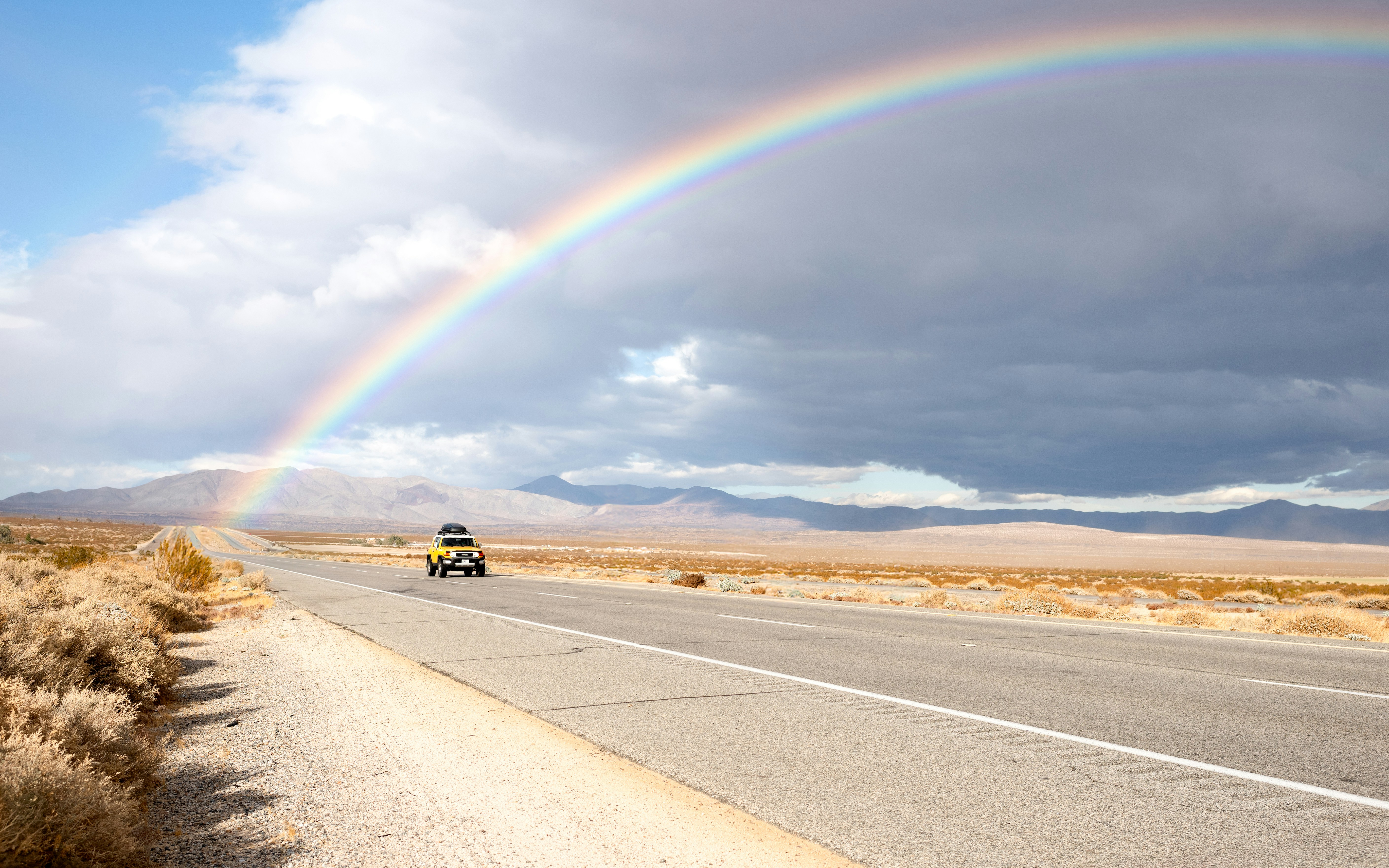 A rainbow over a desert road with a vehicle.