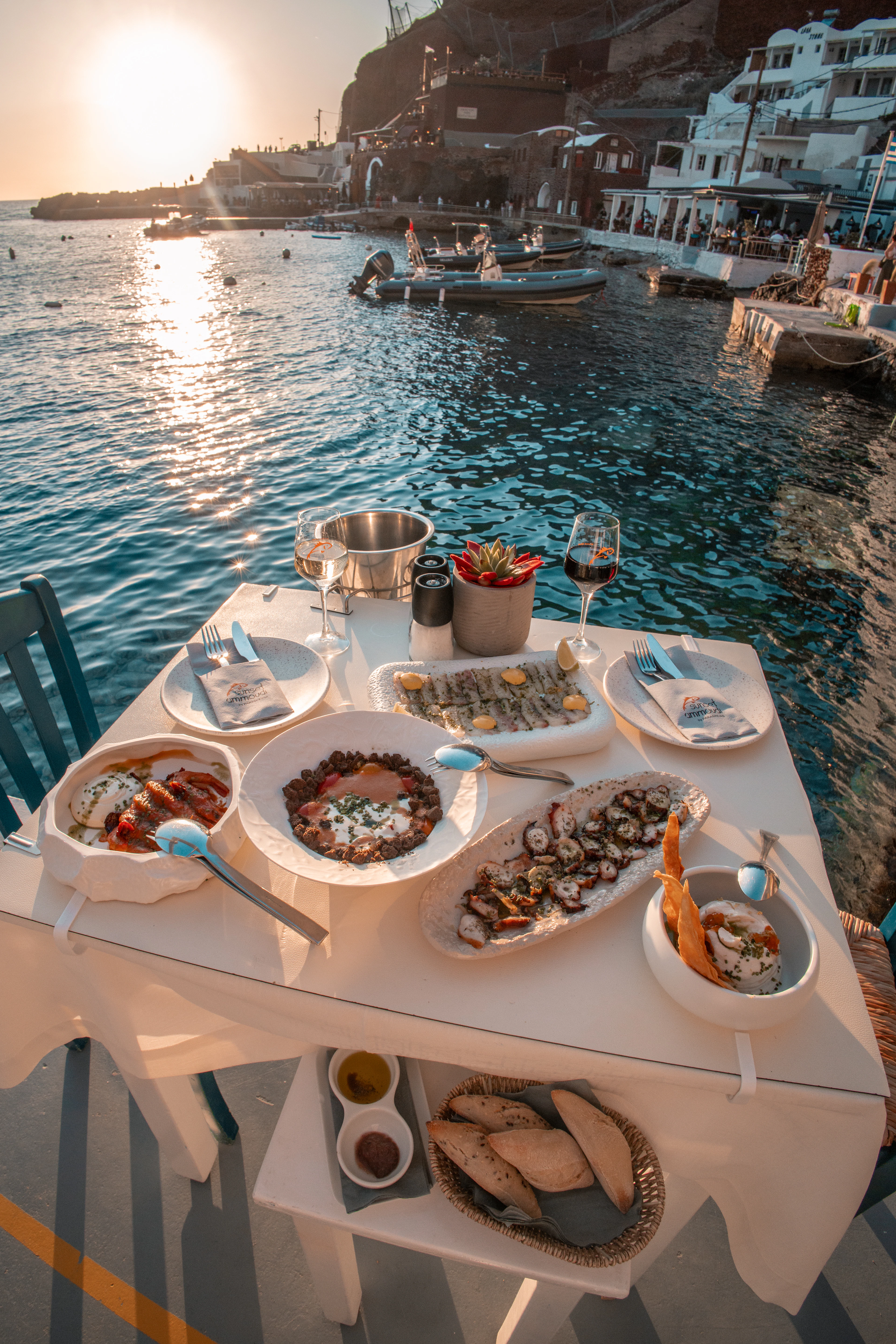 A taverna dinner table set with Greek meze, grilled seafood and a bread basket at the water's edge in Fira, Santorini, at sunset.