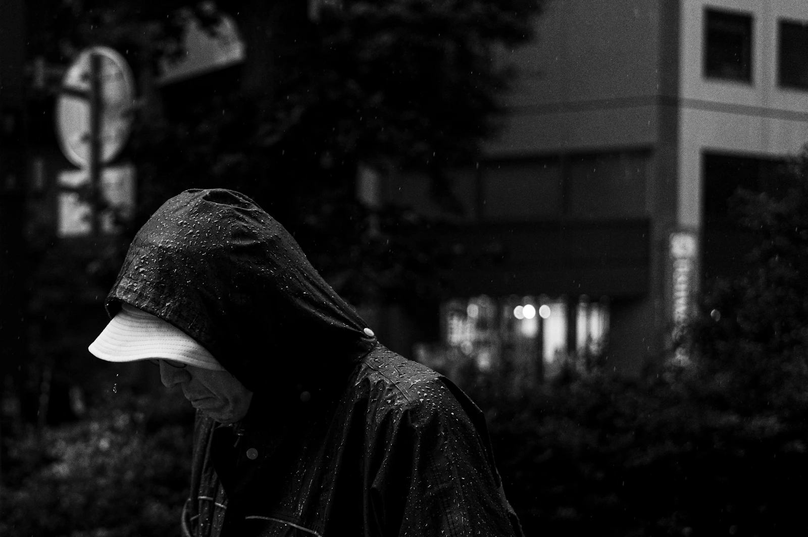 A figure in a hooded raincoat walking through a rain-soaked city street at dusk