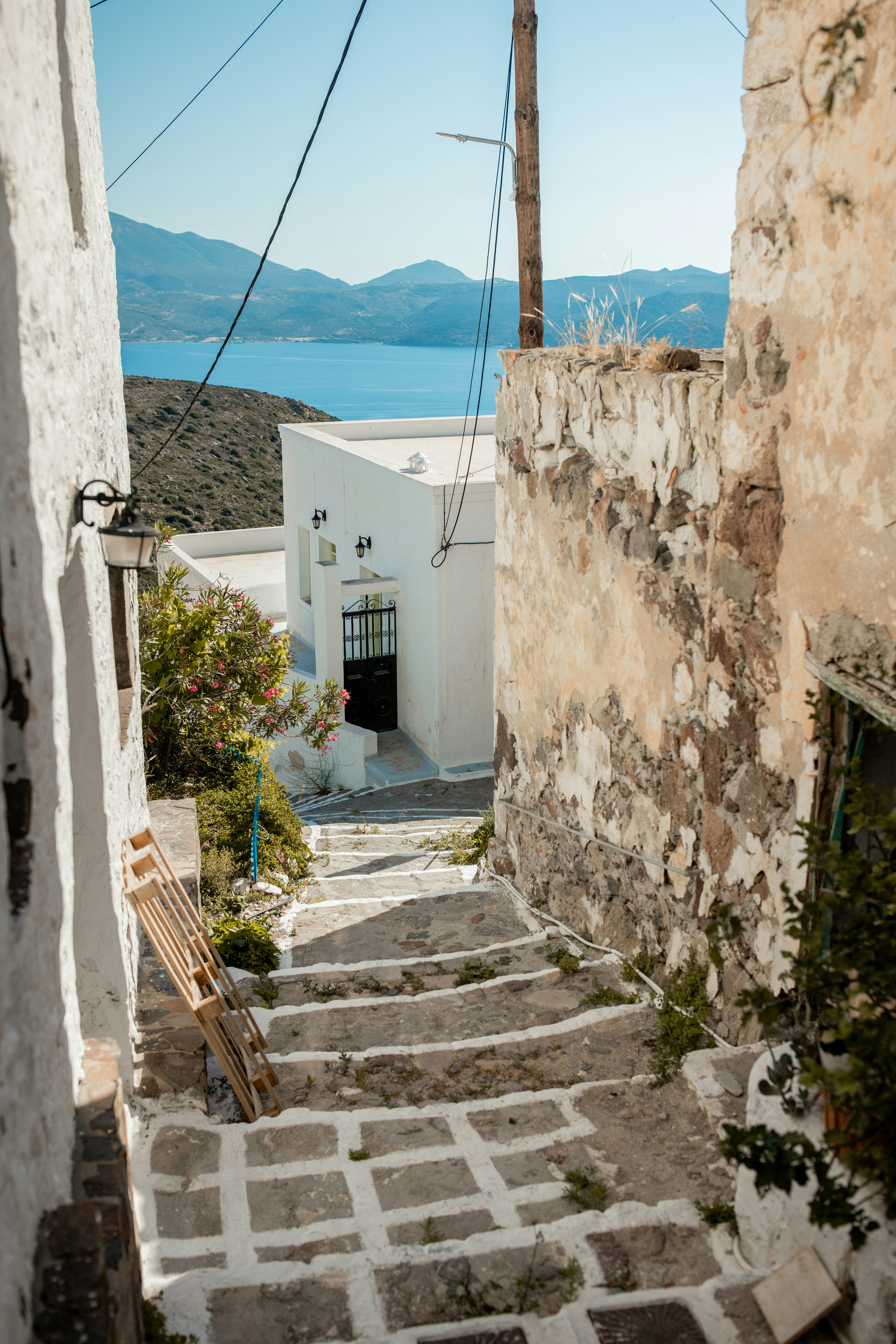 A stepped stone lane between a whitewashed house and a dry-stone wall, dropping past an oleander toward the hazy blue Aegean and the mountains beyond.