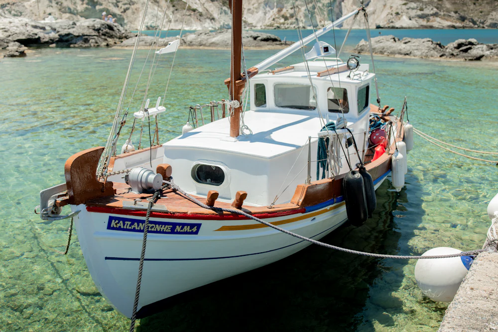 A traditional Greek wooden boat with a small white wheelhouse lying in shallow clear turquoise water off a rocky shore.