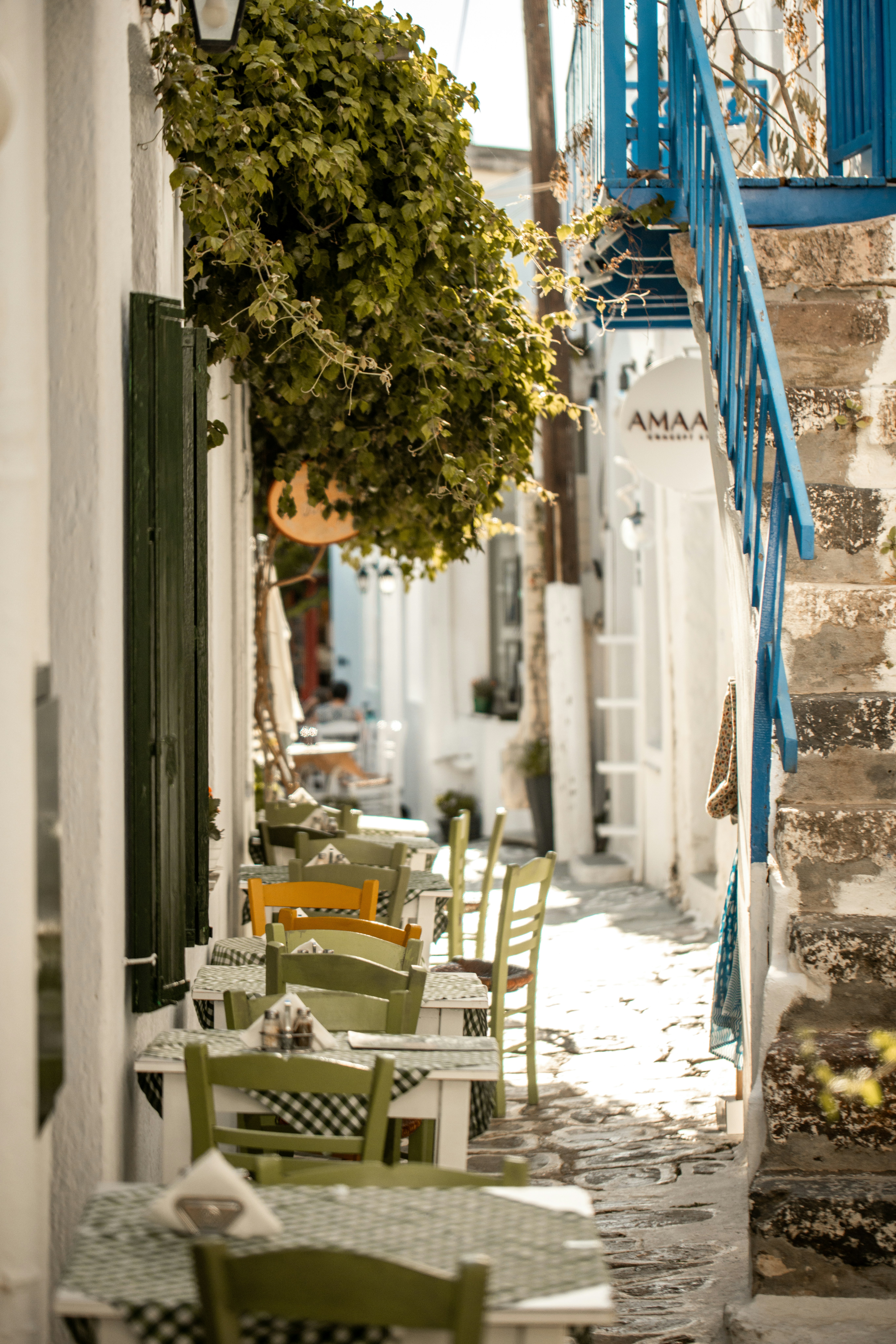 A narrow whitewashed alley with wooden chairs and small tables set out under a vine, deep shade on one side and hard sun on the other.
