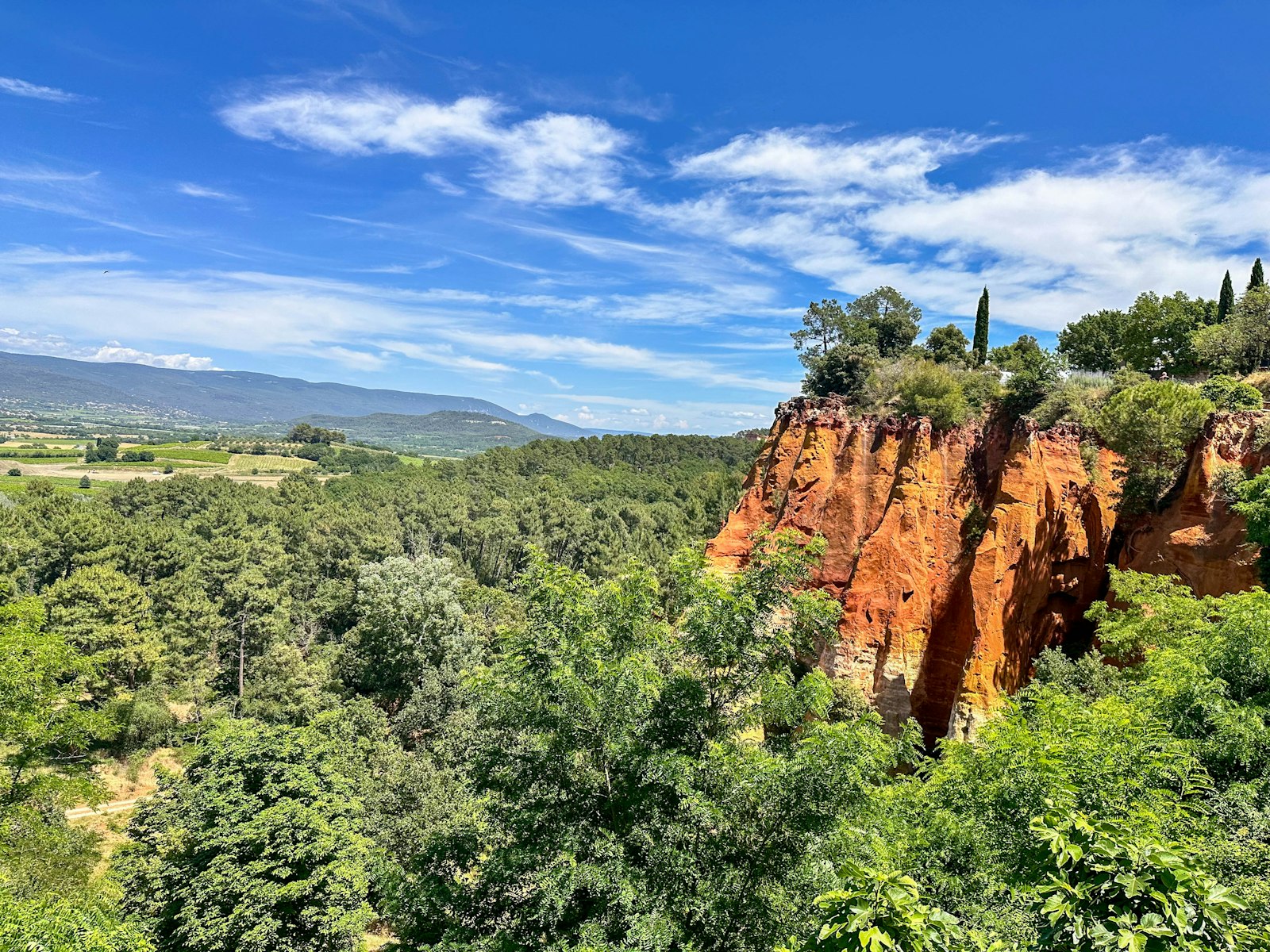 Red and orange ochre cliffs rising out of green pine forest at Roussillon, with the Luberon hills on the horizon