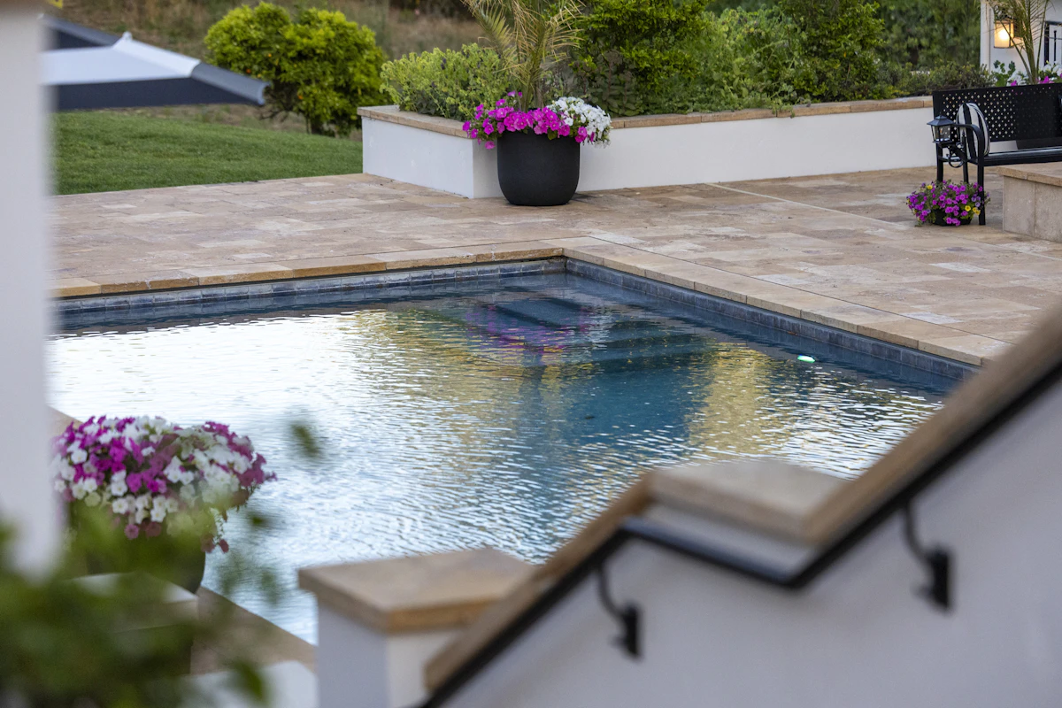 Une piscine rectangulaire bordée d'une terrasse en pierre claire, avec des massifs de fleurs roses et un banc en fer forgé au fond du jardin