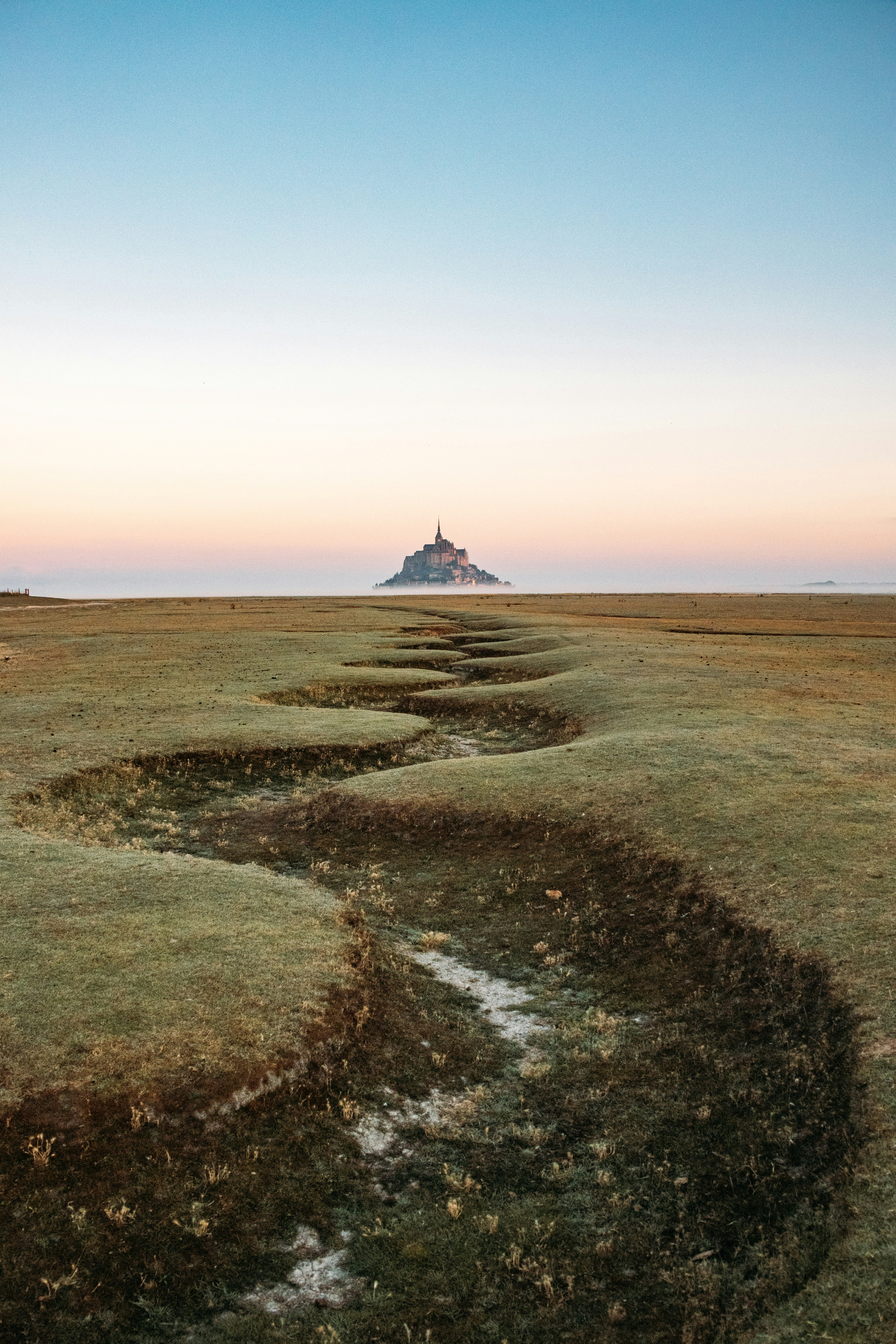 Mont saint-michel across a winding grassy plain at dawn.