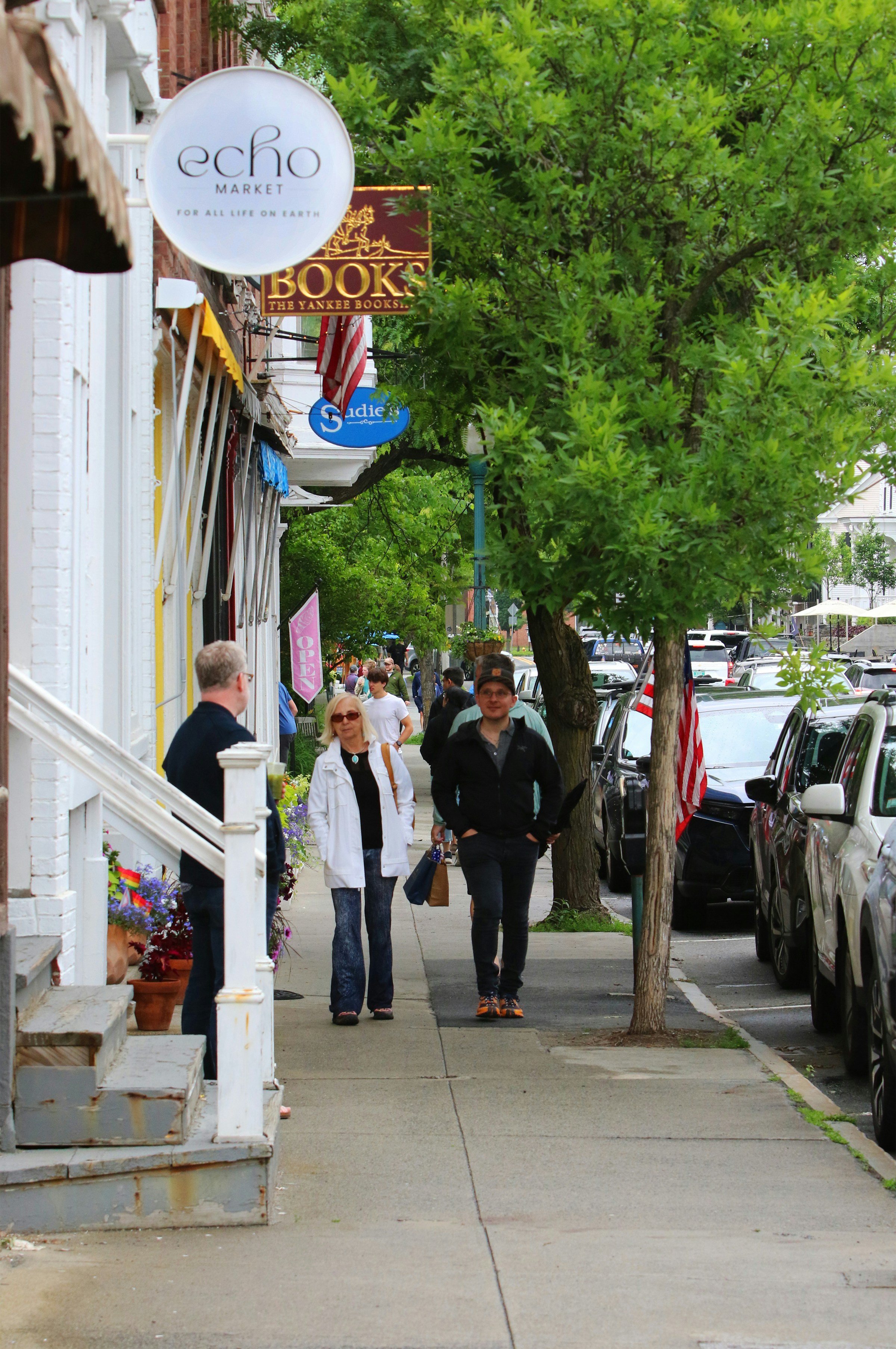 People walking on a sidewalk past shops and trees.