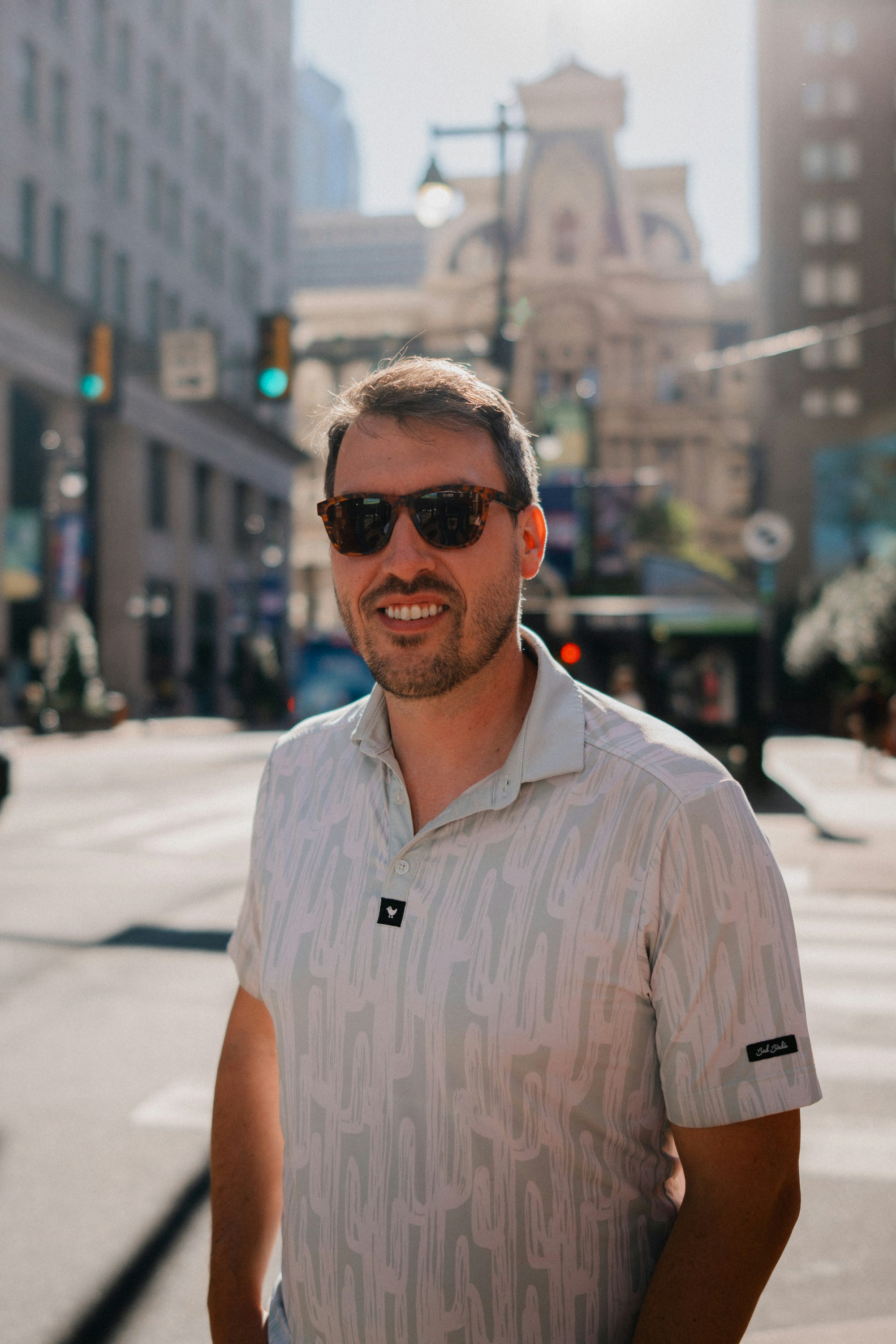 A man in sunglasses smiles on a sunny city street.