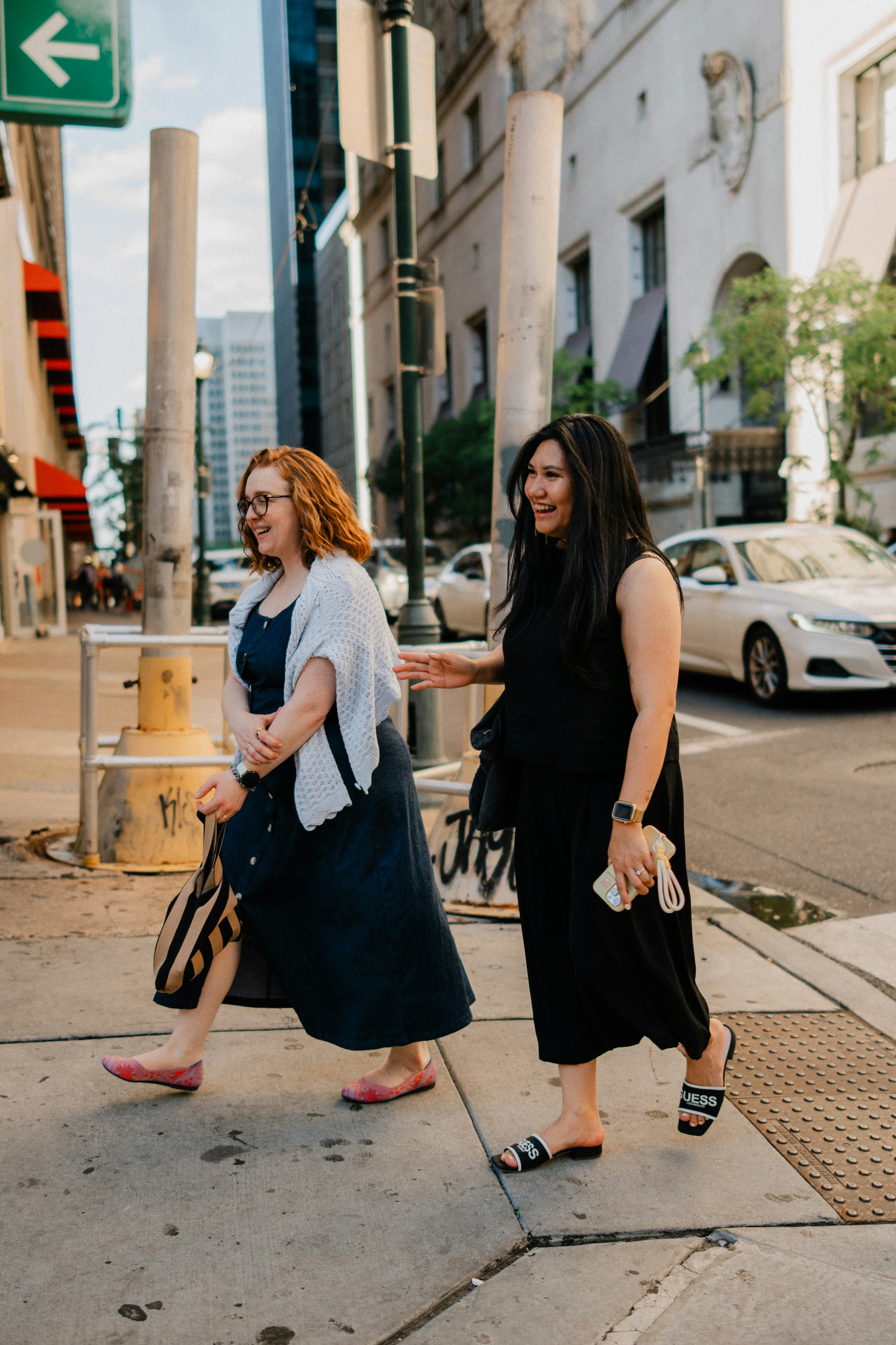Two smiling women walk on a city sidewalk.
