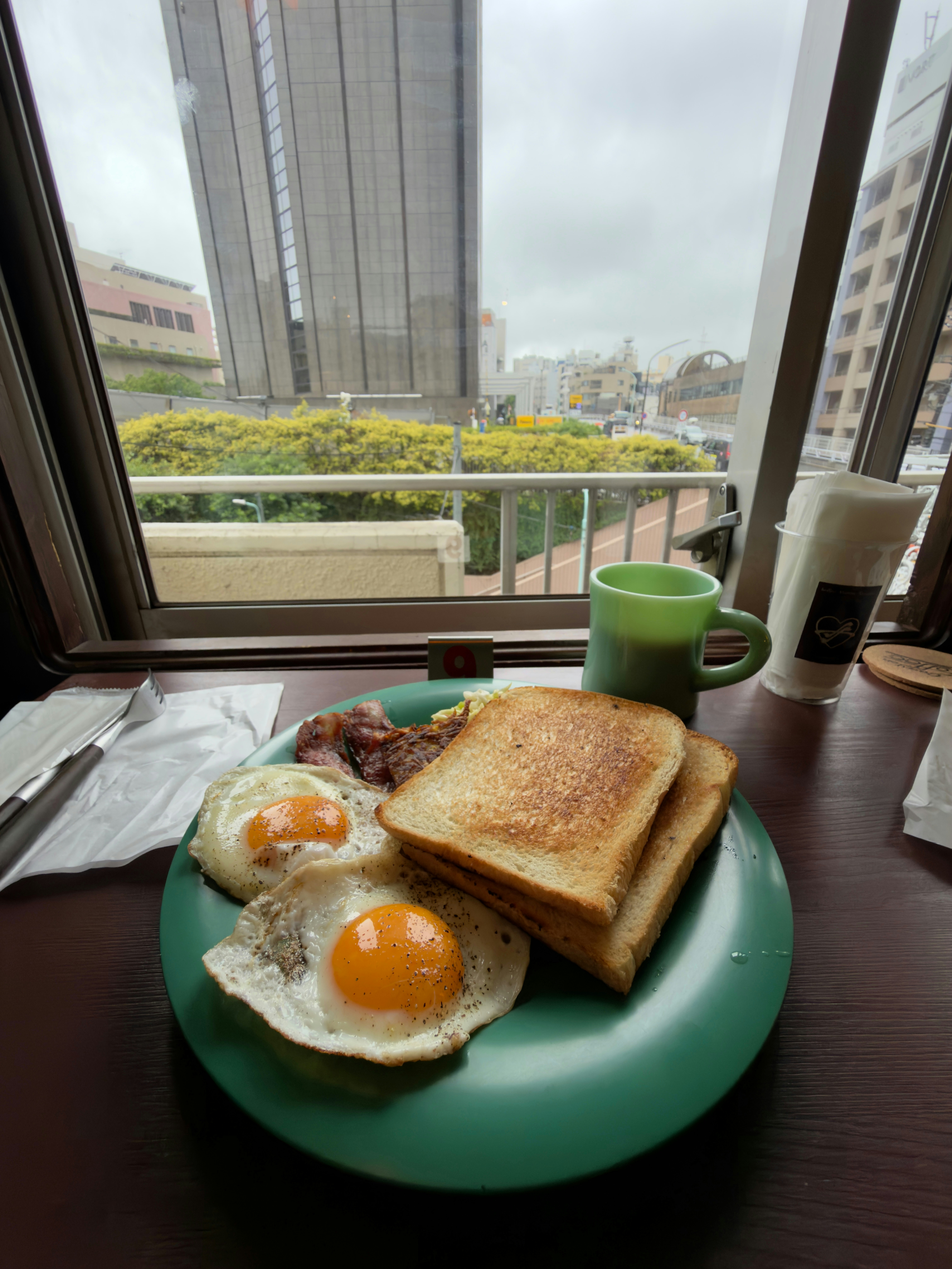 Desayuno con huevos, tostadas y café junto a una ventana urbana.