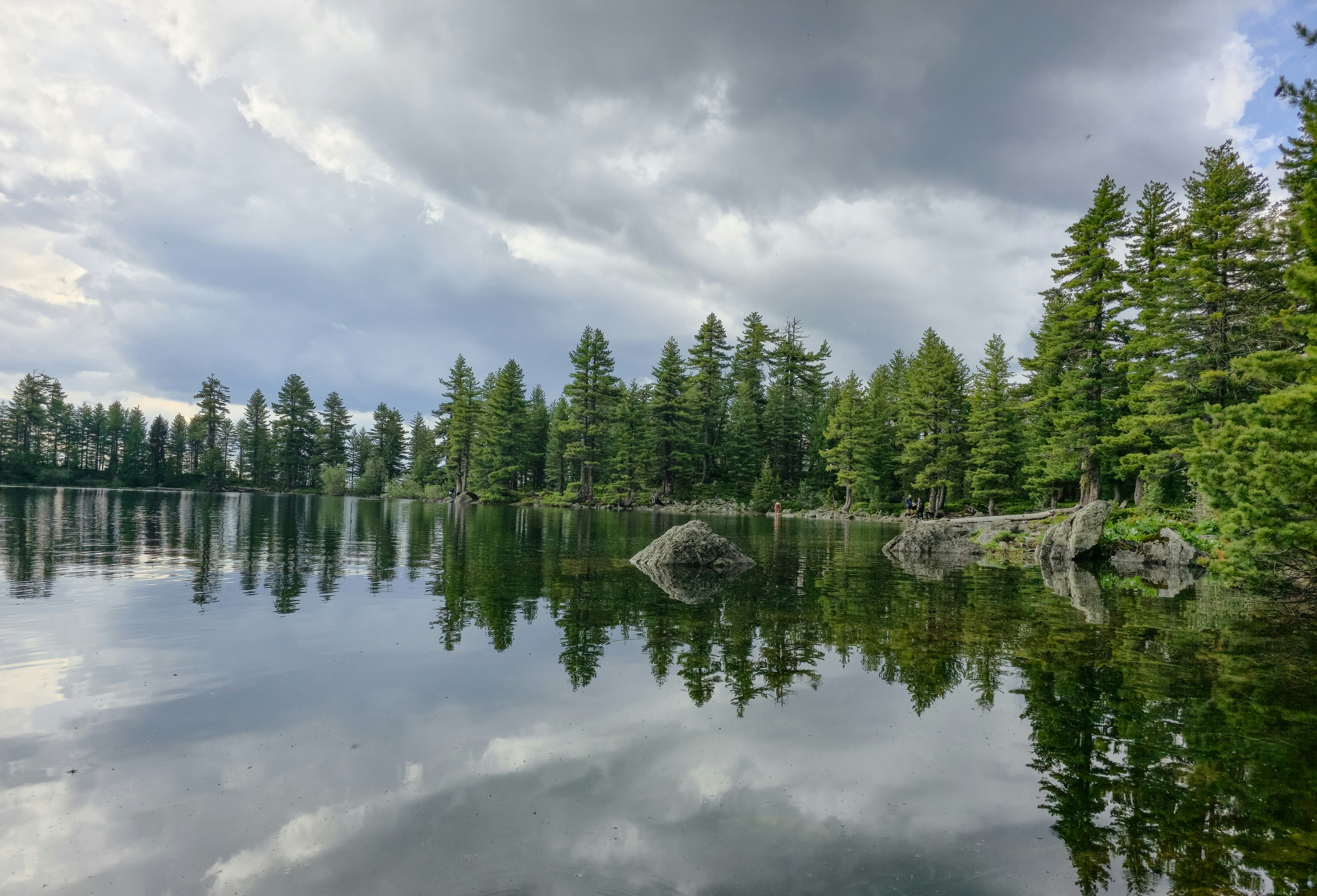 A calm lake reflecting evergreen trees and cloudy sky.