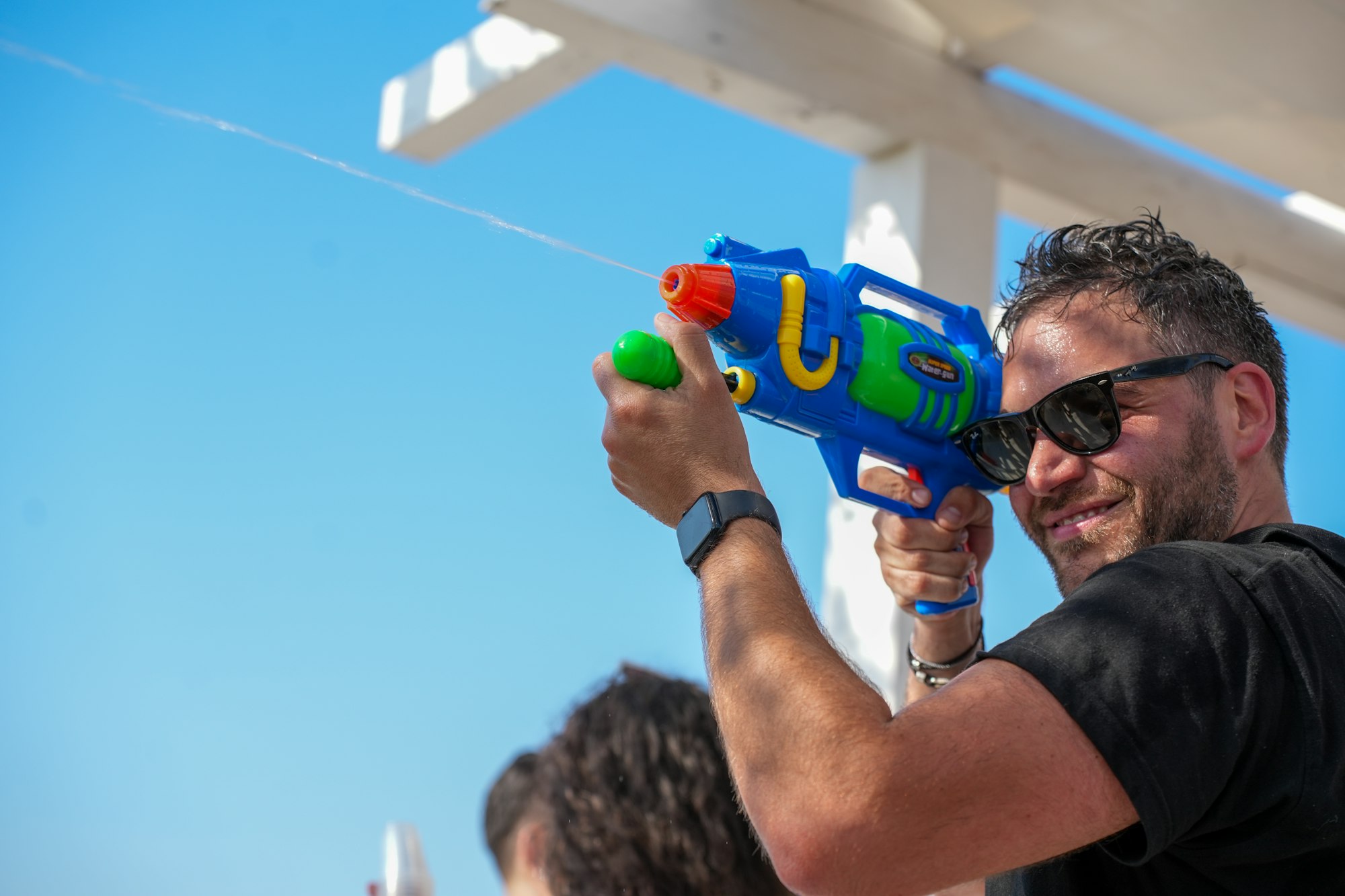 Smiling man shoots water gun under clear blue sky.