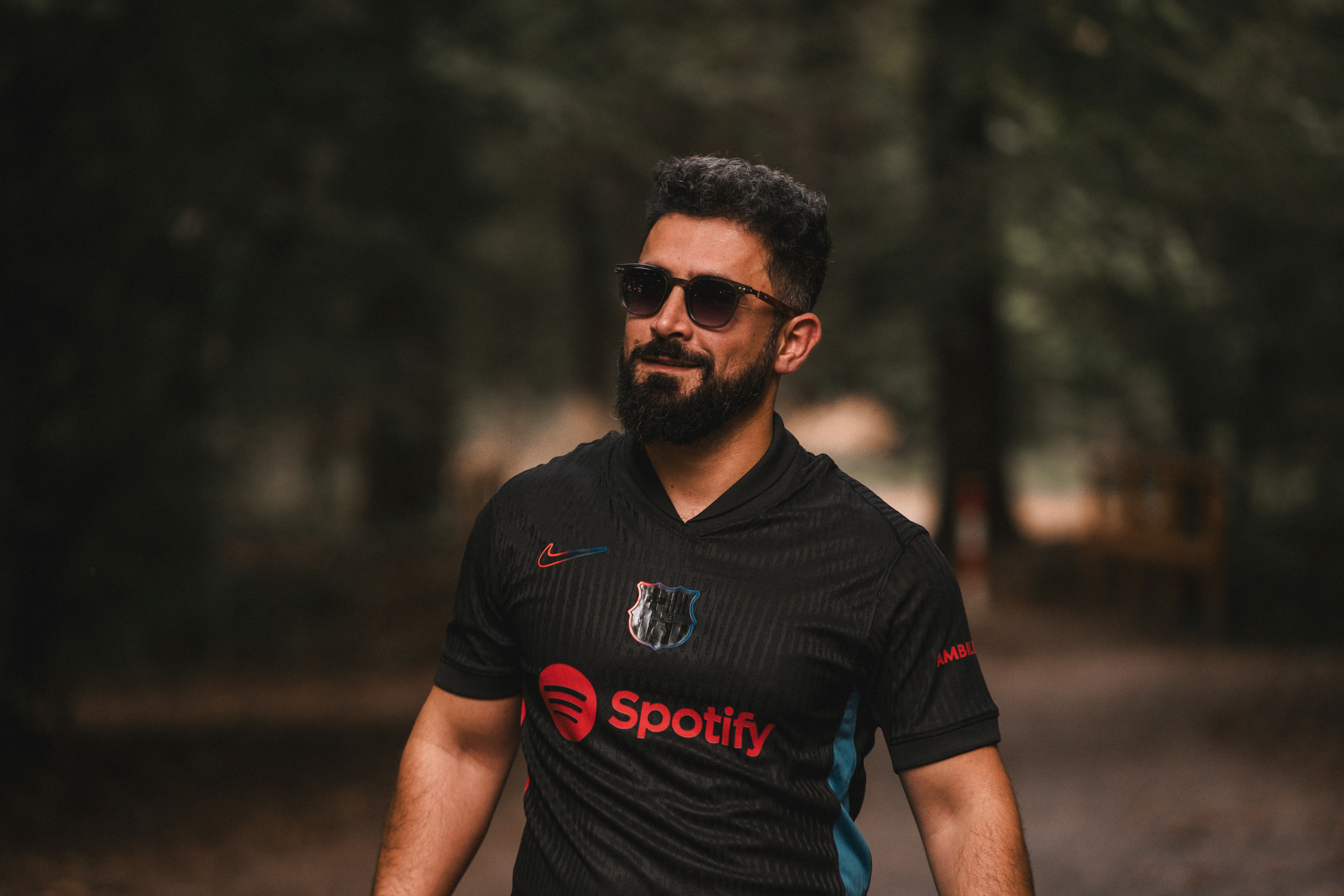 Uomo barbuto con occhiali da sole e maglietta spotify in una foresta.