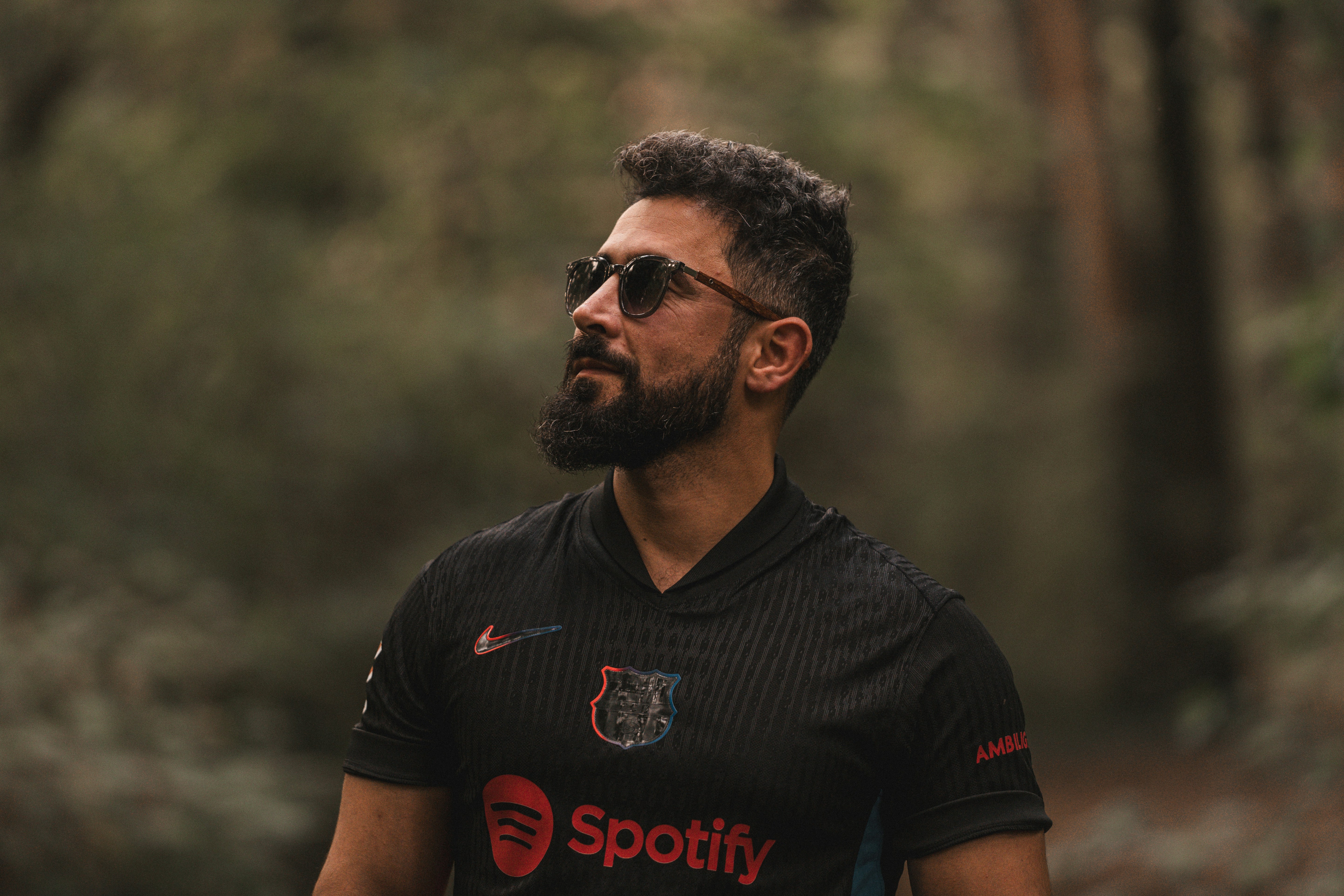 Uomo barbuto con occhiali da sole e maglia in una foresta.
