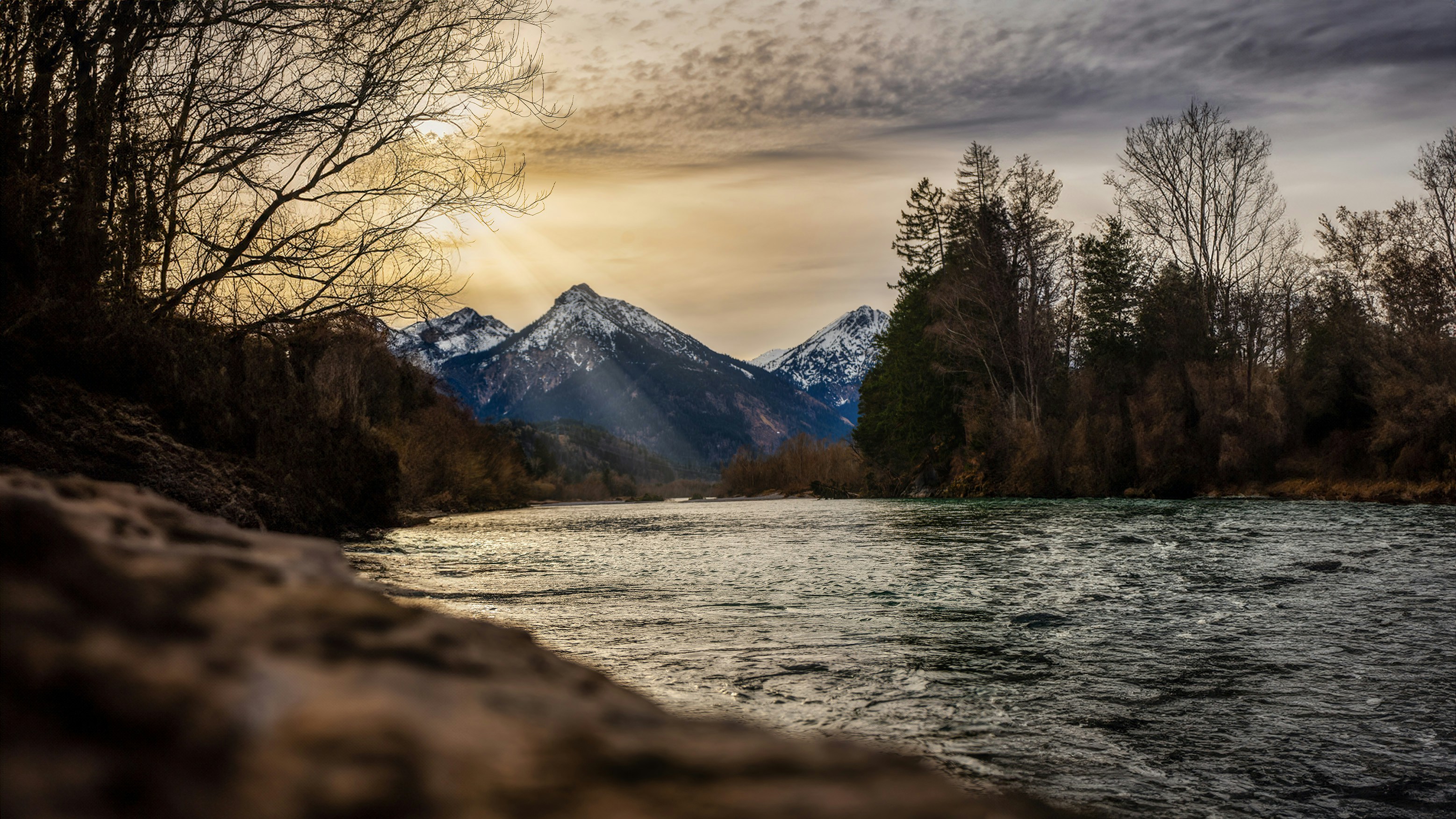 A river flows towards snow-capped mountains under a dramatic sky