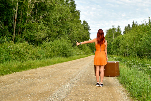 Red-haired woman in orange dress hitchhiking with a suitcase.