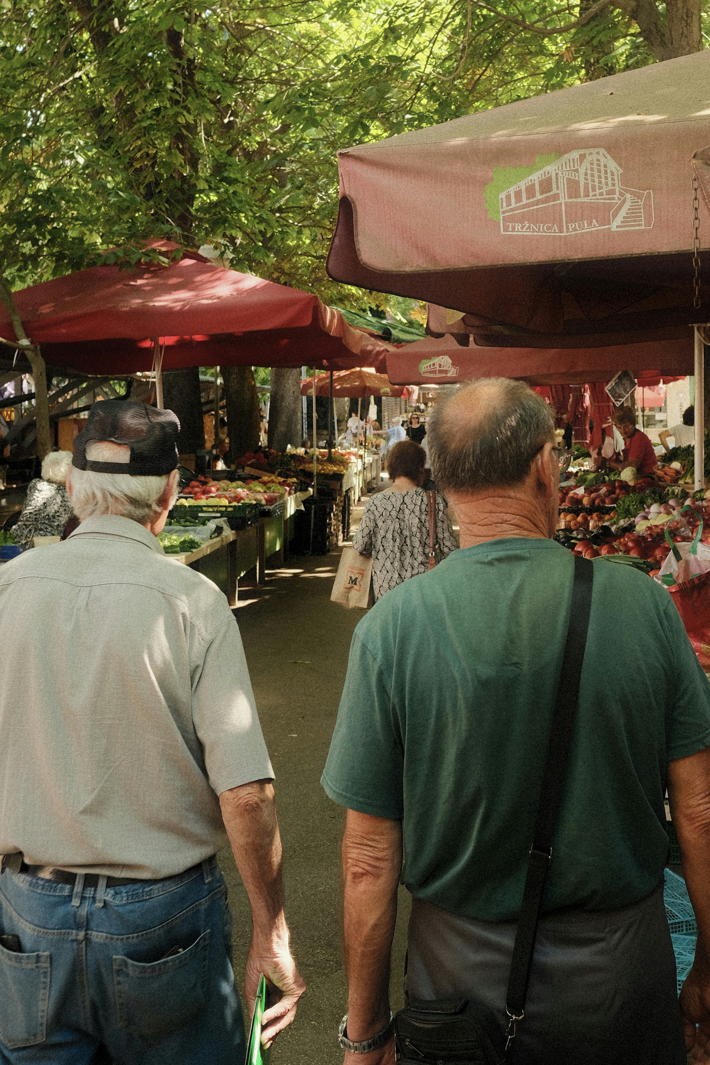 Two men walk through a bustling outdoor market with stalls.