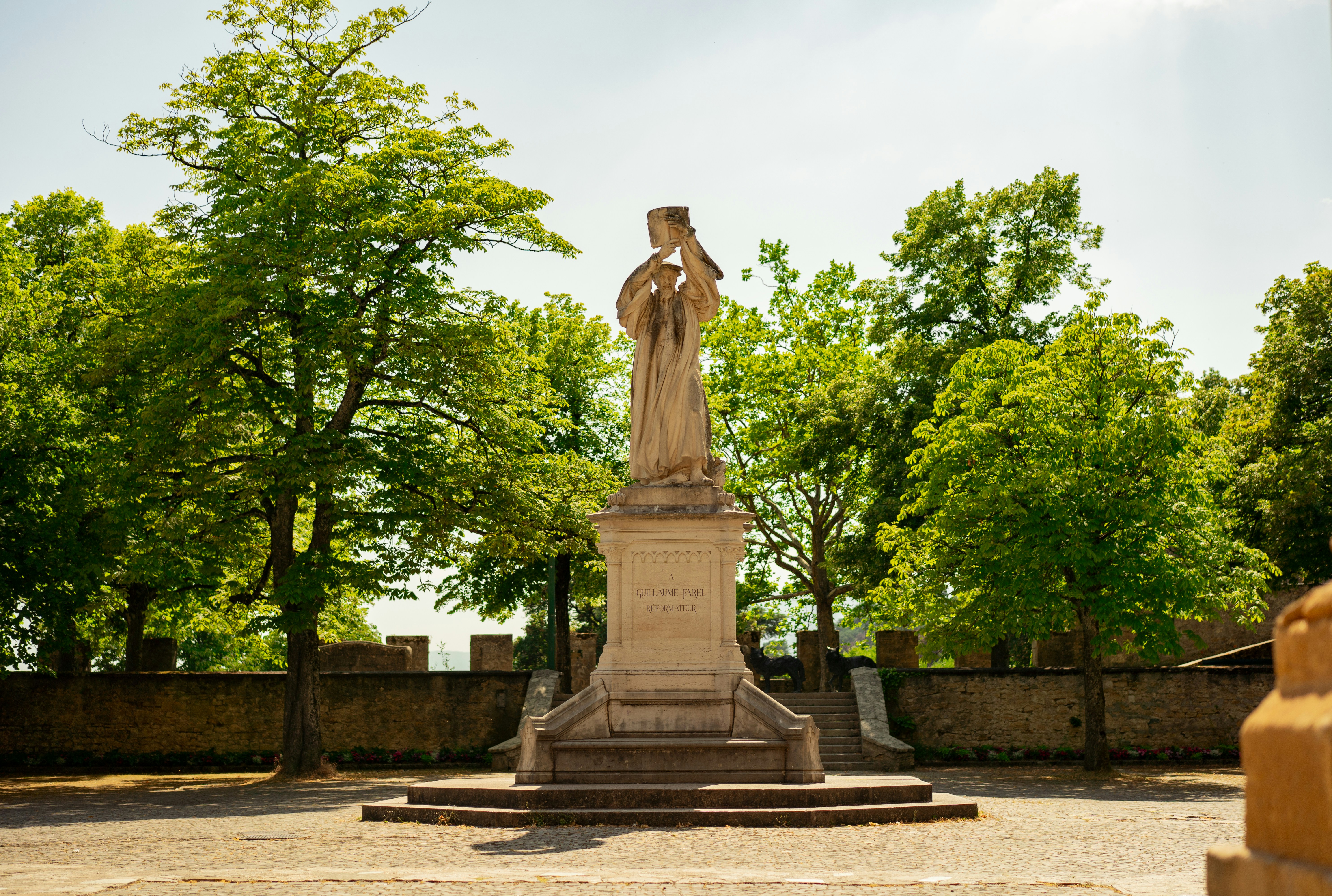 A stone statue stands among green trees and a wall.