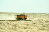 An orange dump truck drives through a dusty, sandy landscape