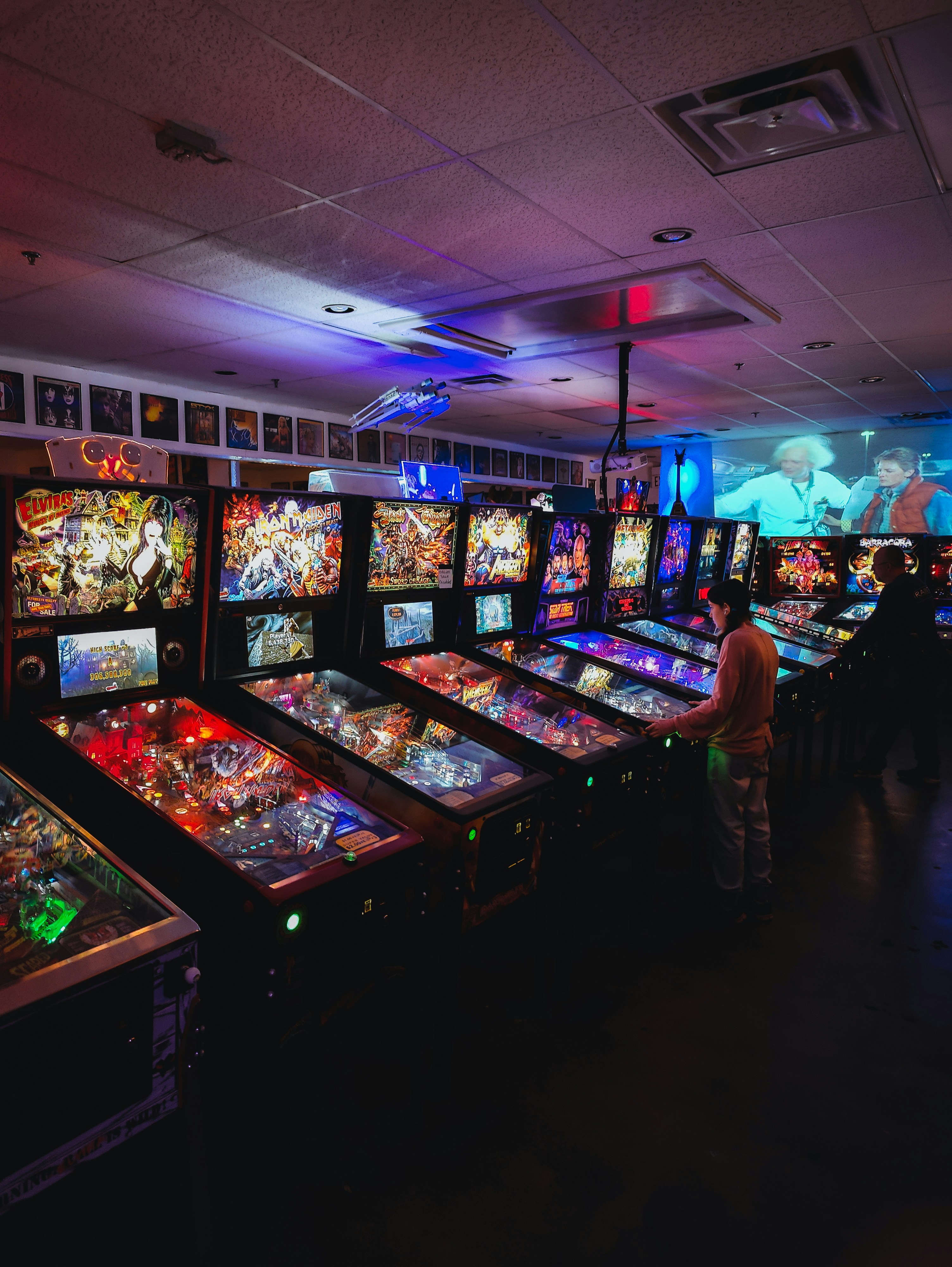 Pessoas jogando máquinas de pinball coloridas em um fliperama mal iluminado.