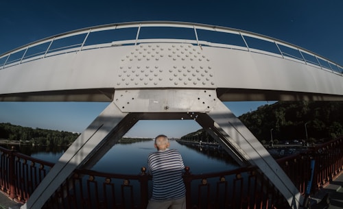Man in striped shirt stands on bridge overlooking river.