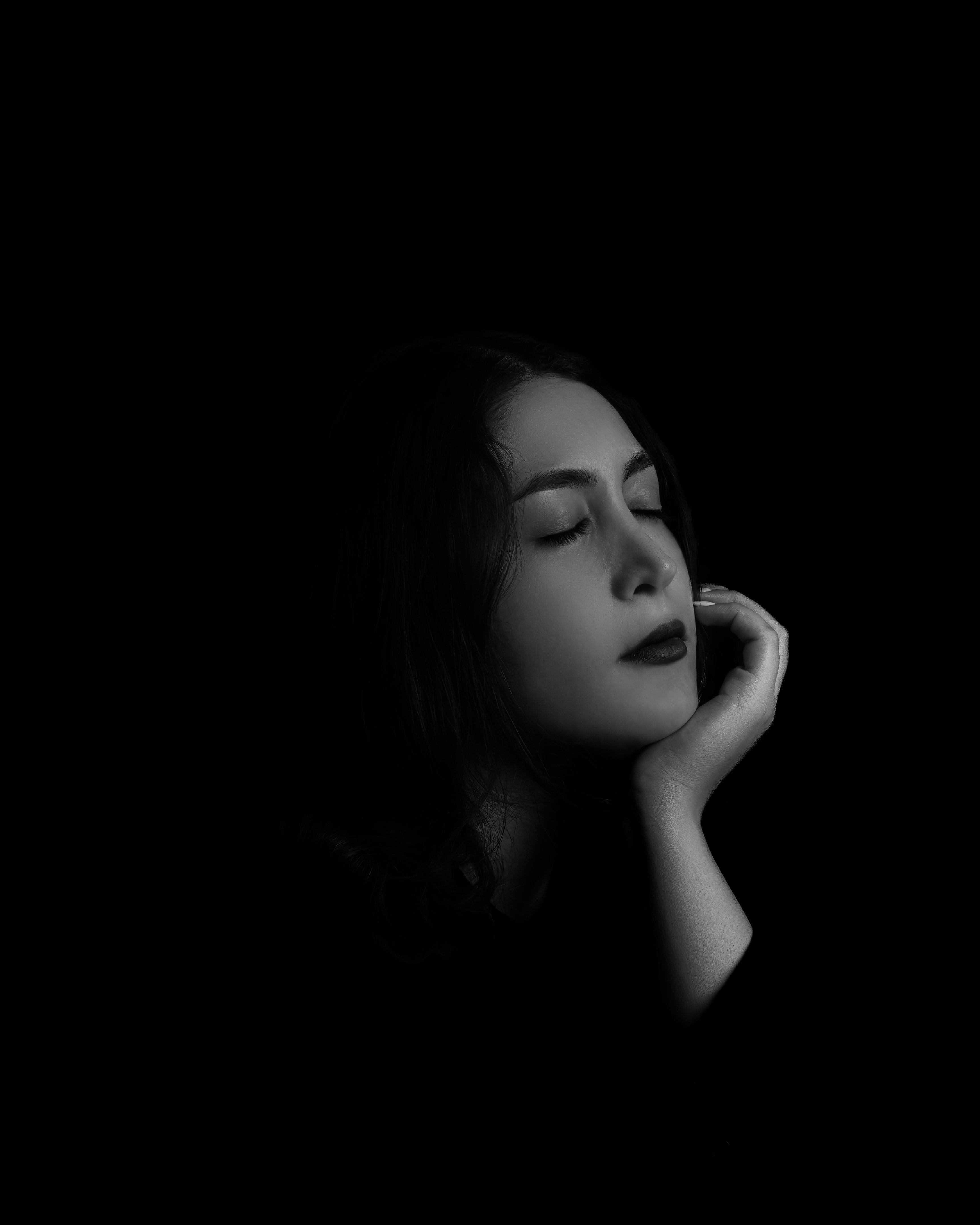 A black and white three-quarter portrait of a woman resting her hand beneath her chin. Her head is slightly raised while soft lighting and selective focus emphasize facial features and texture.