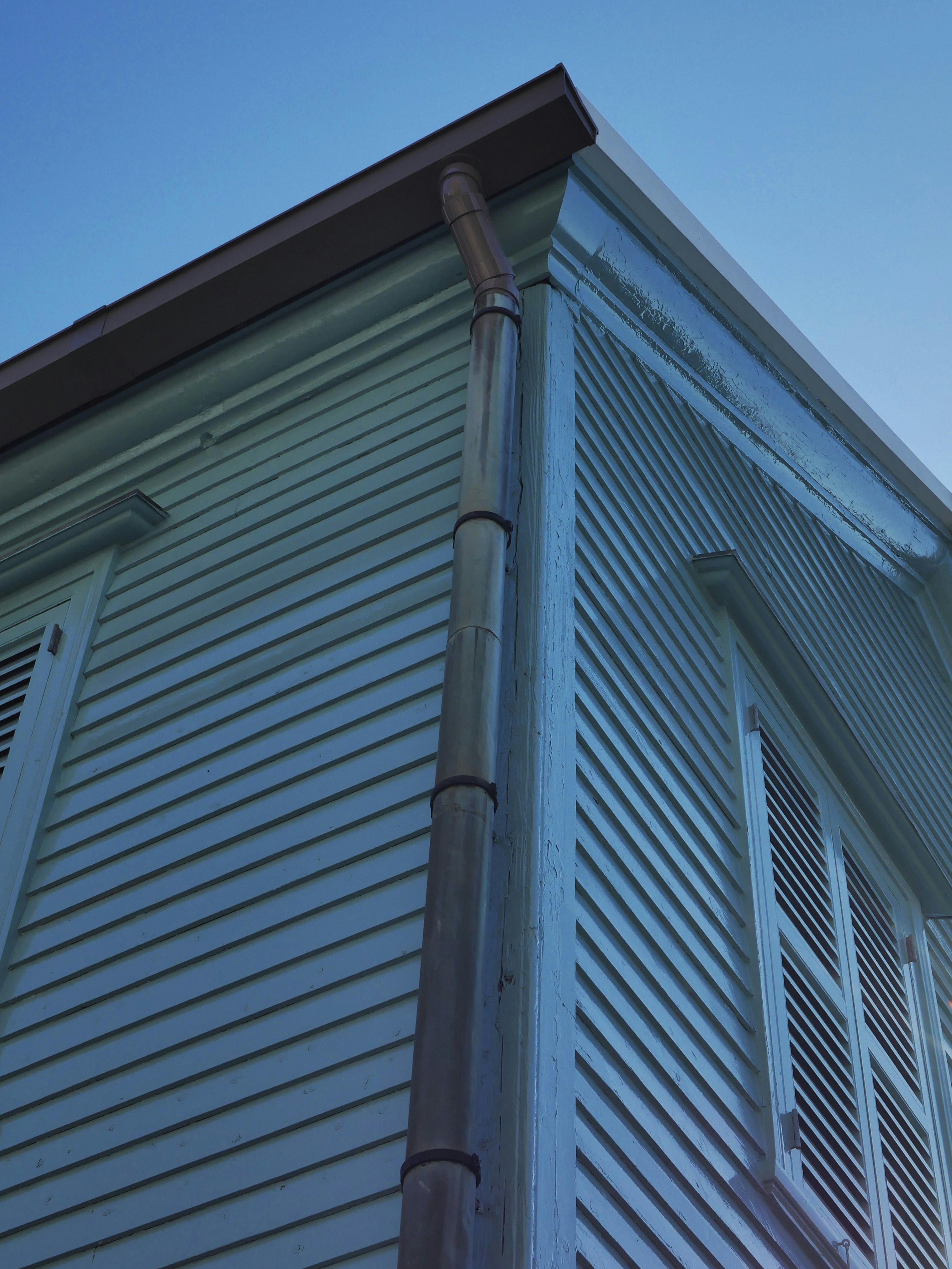 Light-colored building corner with shutters, downspout, and blue sky.
