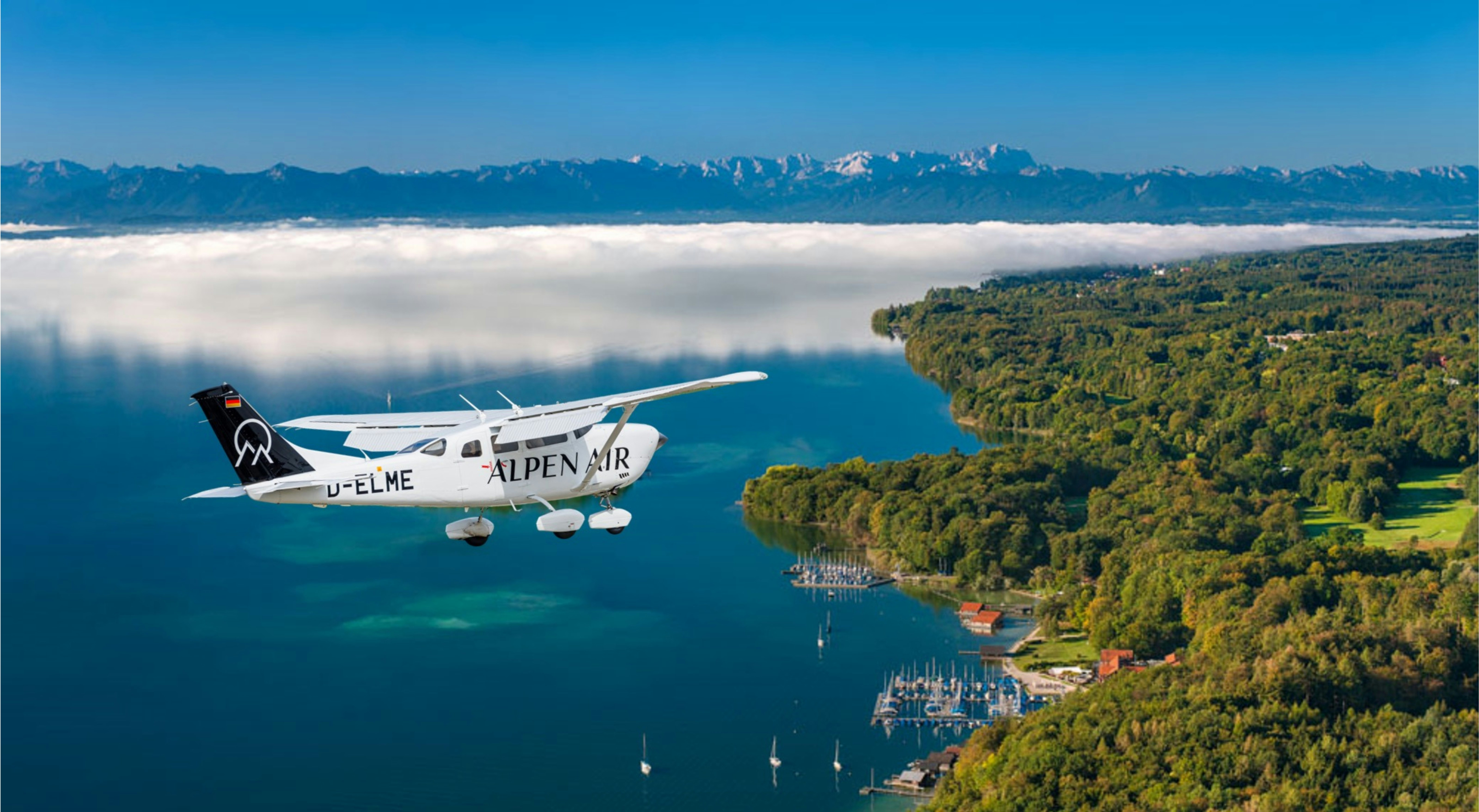 White plane flying over a lake, green land, and mountains.