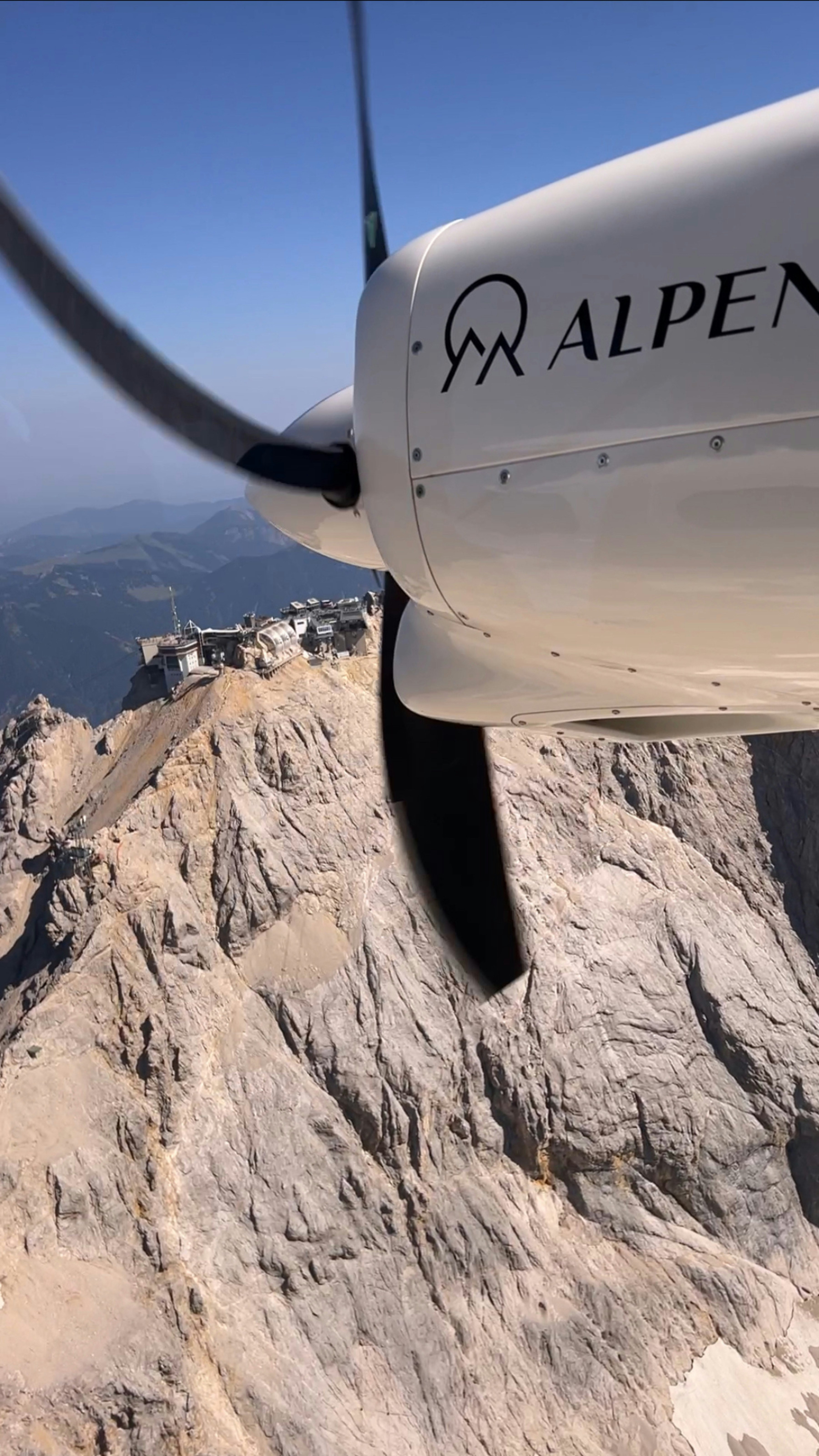 Airplane propeller above a rocky mountain peak with buildings.