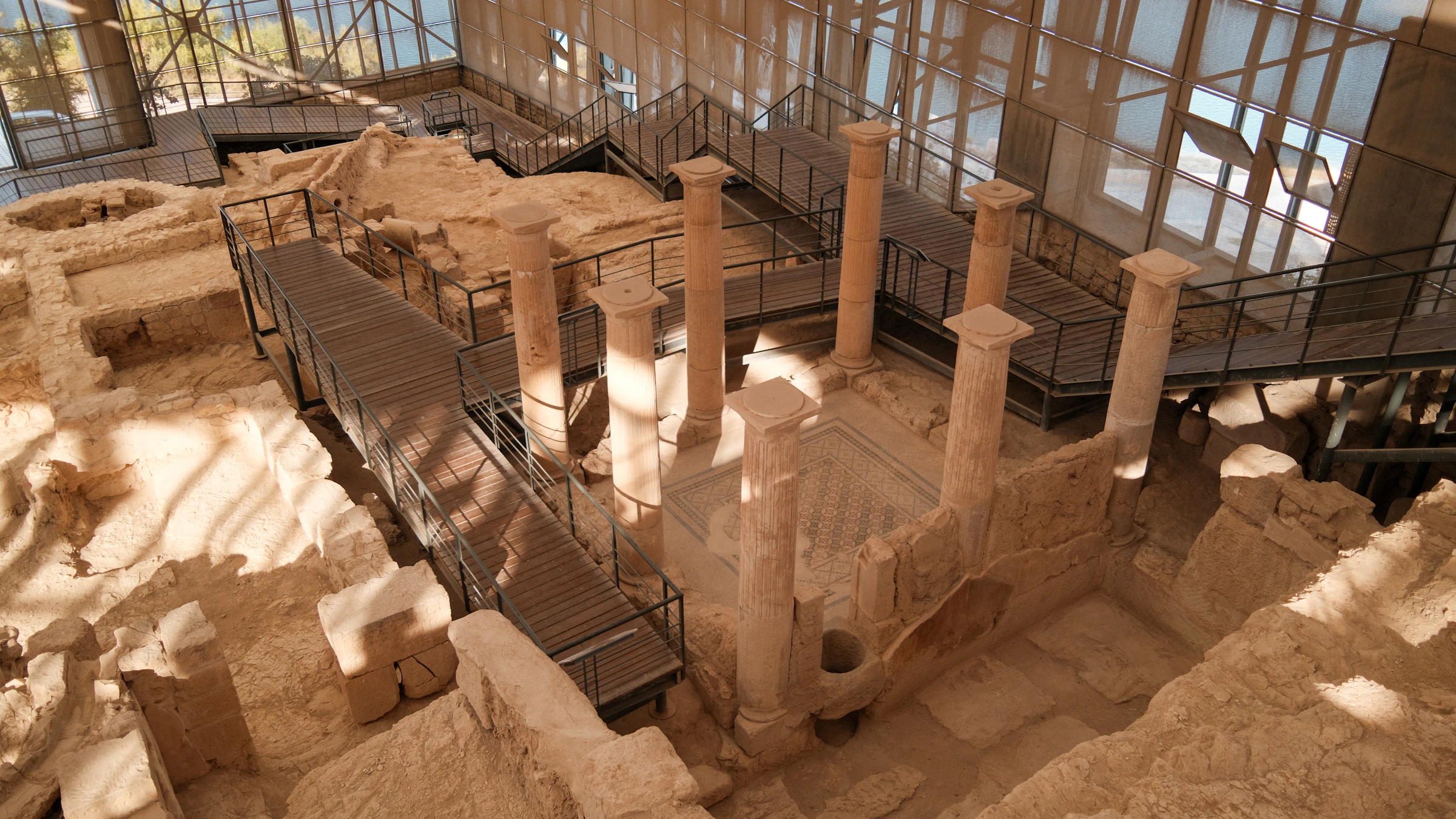 An excavated Roman villa with standing columns and a patterned mosaic floor, sheltered under a modern protective roof with a raised visitor walkway