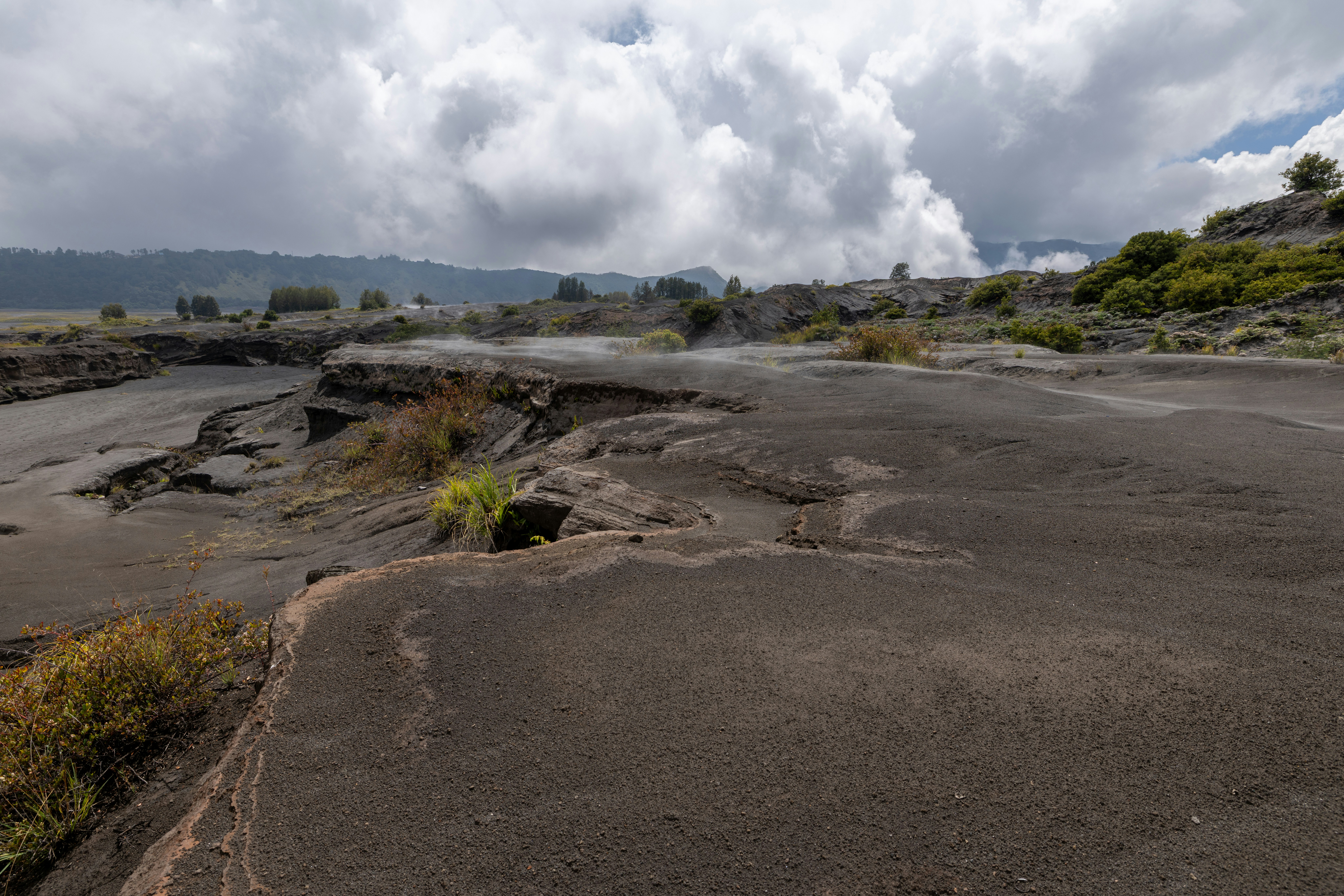 Volcanic landscape with dark ash, steam, and cloudy sky.