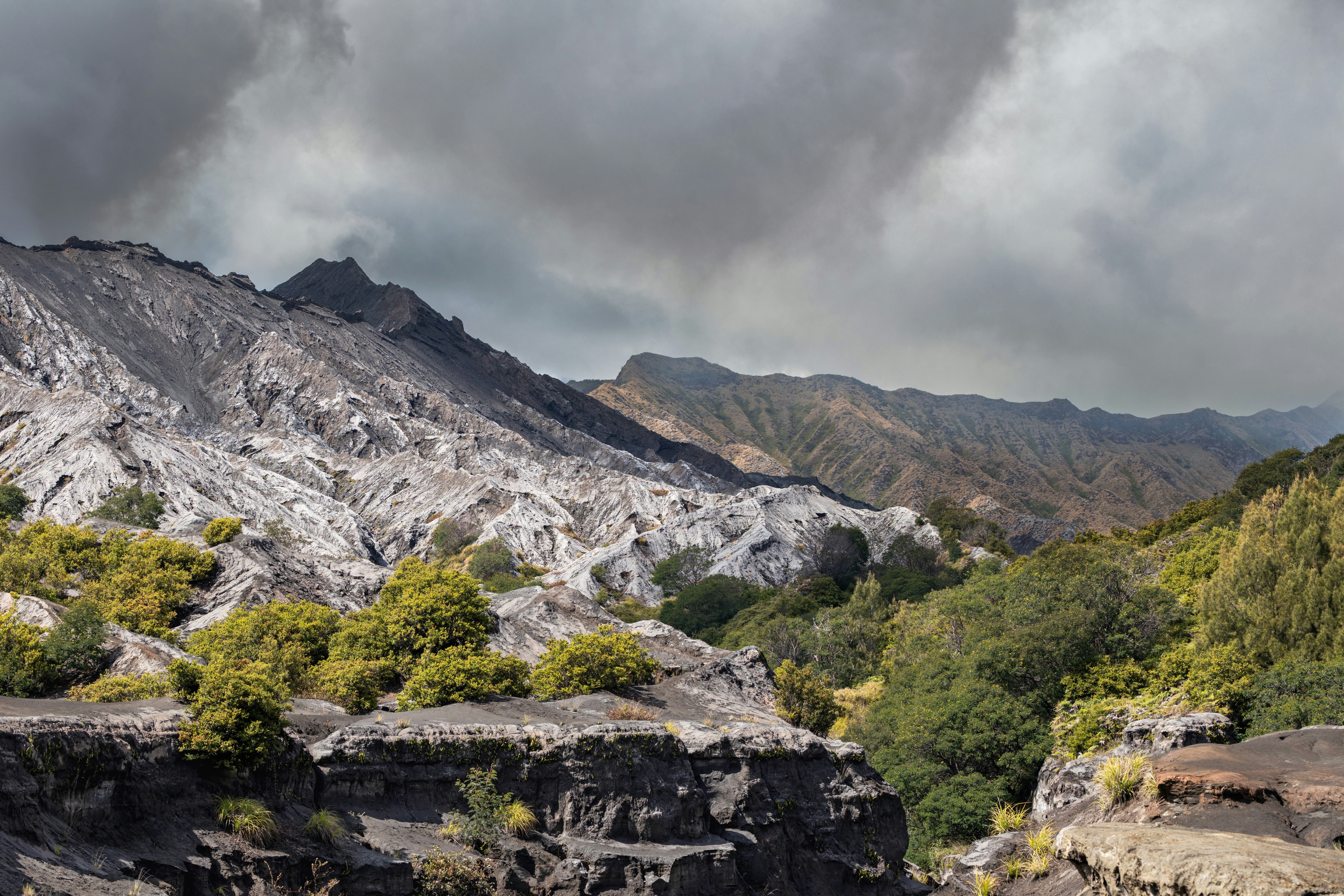 Ash-covered volcanic mountains and green vegetation under a cloudy sky.
