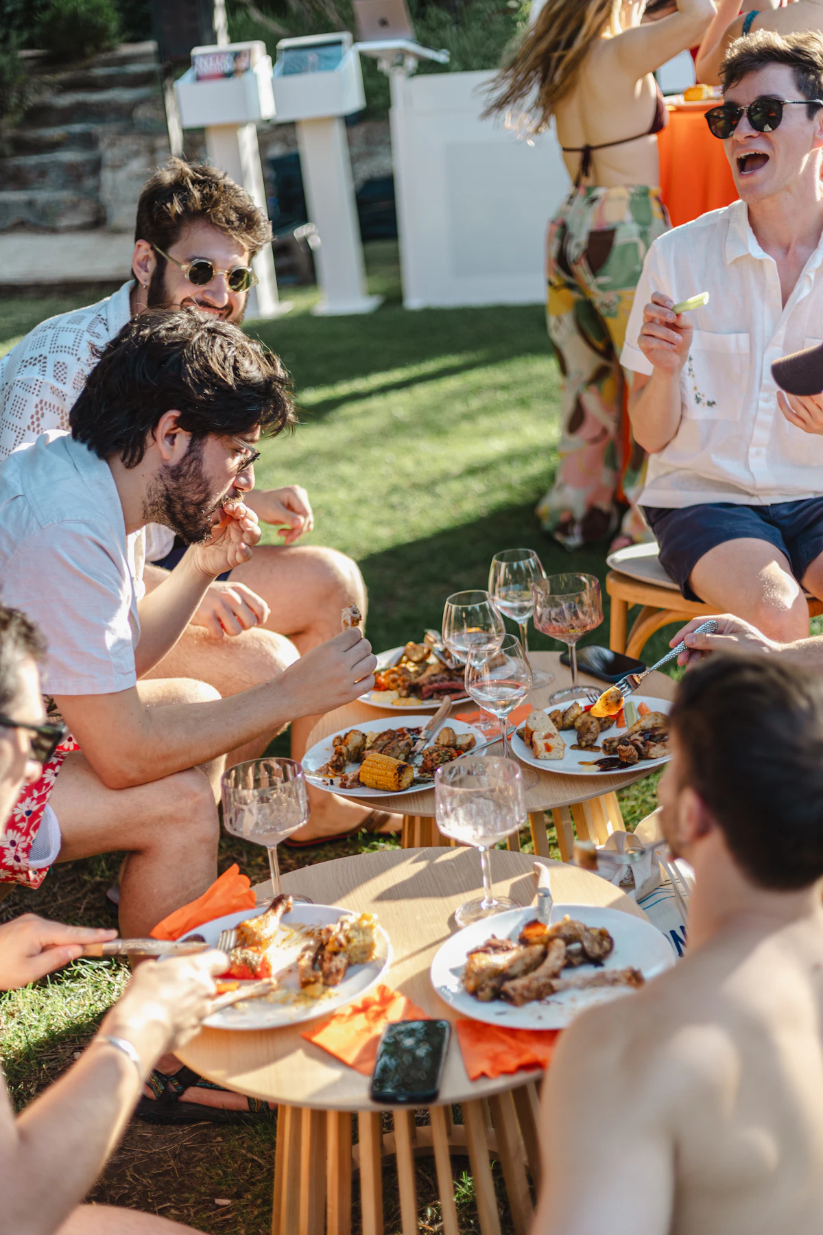 Friends sitting on the grass around a spread of food and open wine on a summer evening