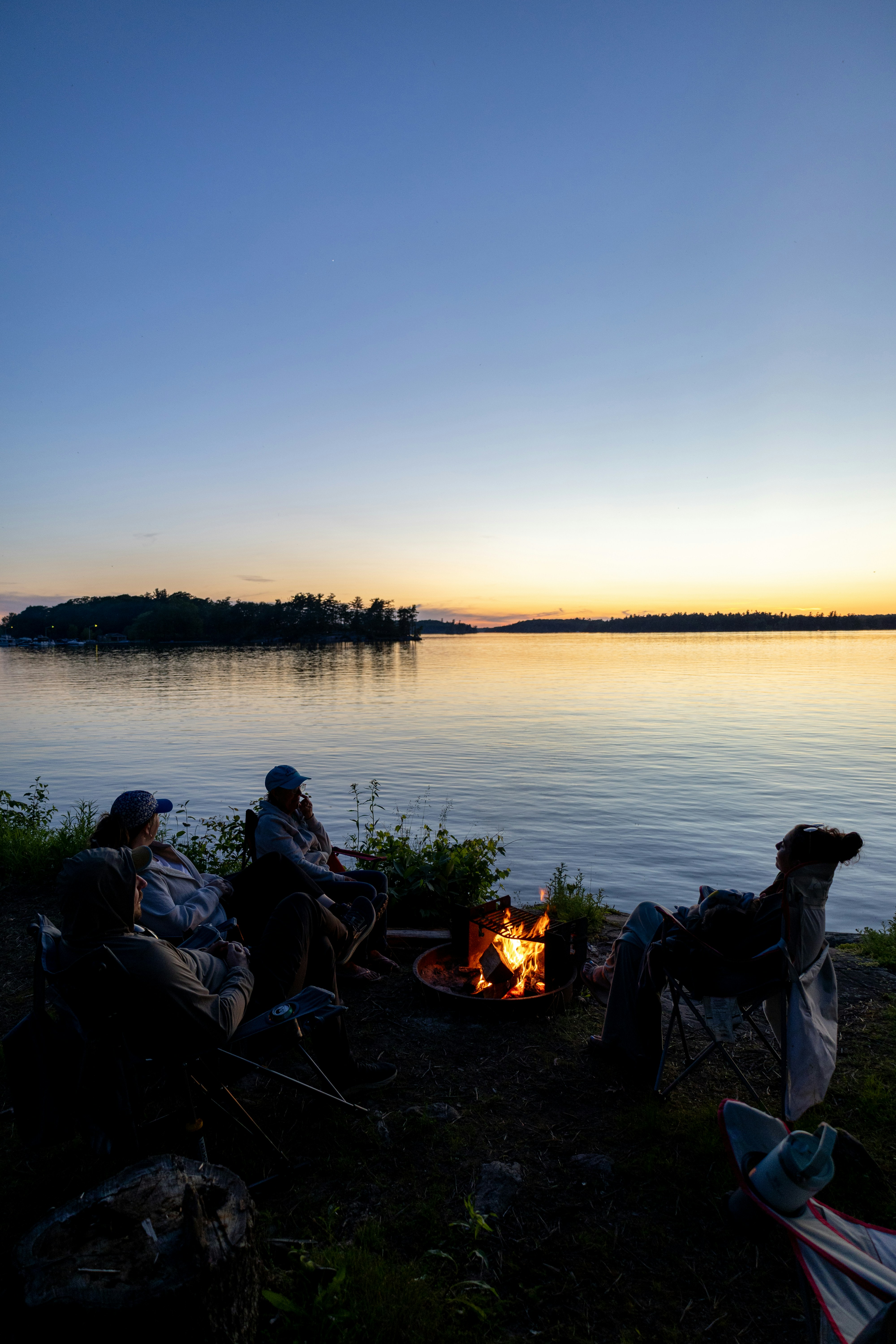 Des gens assis autour d’un feu de camp près d’un lac au coucher du soleil.