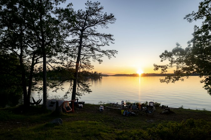 Campsite on a grassy lakeshore at sunset, a small group gathered by their tents under tall pines as the sun sets across the water