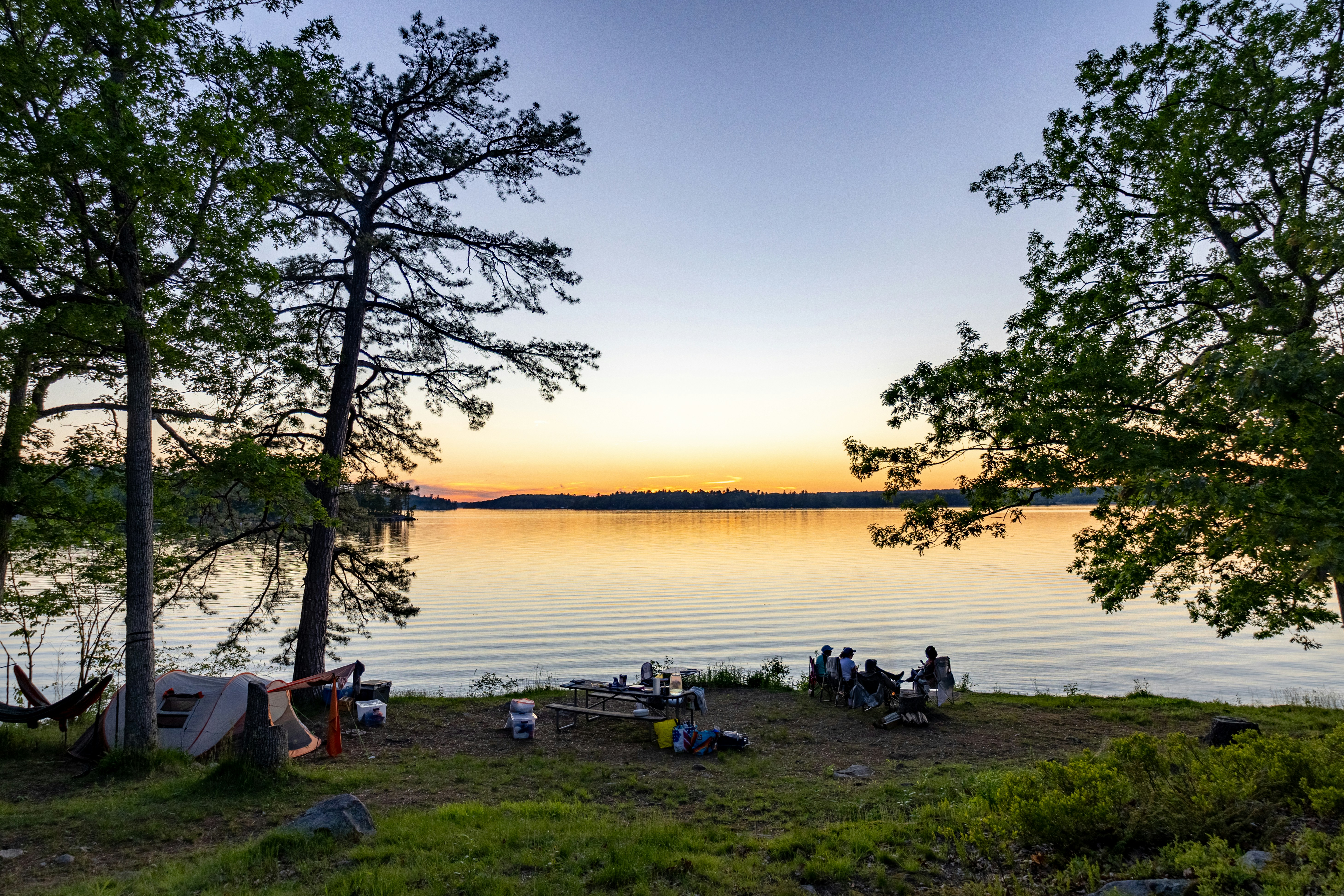 Des gens campant au bord d’un lac au coucher du soleil avec des arbres.