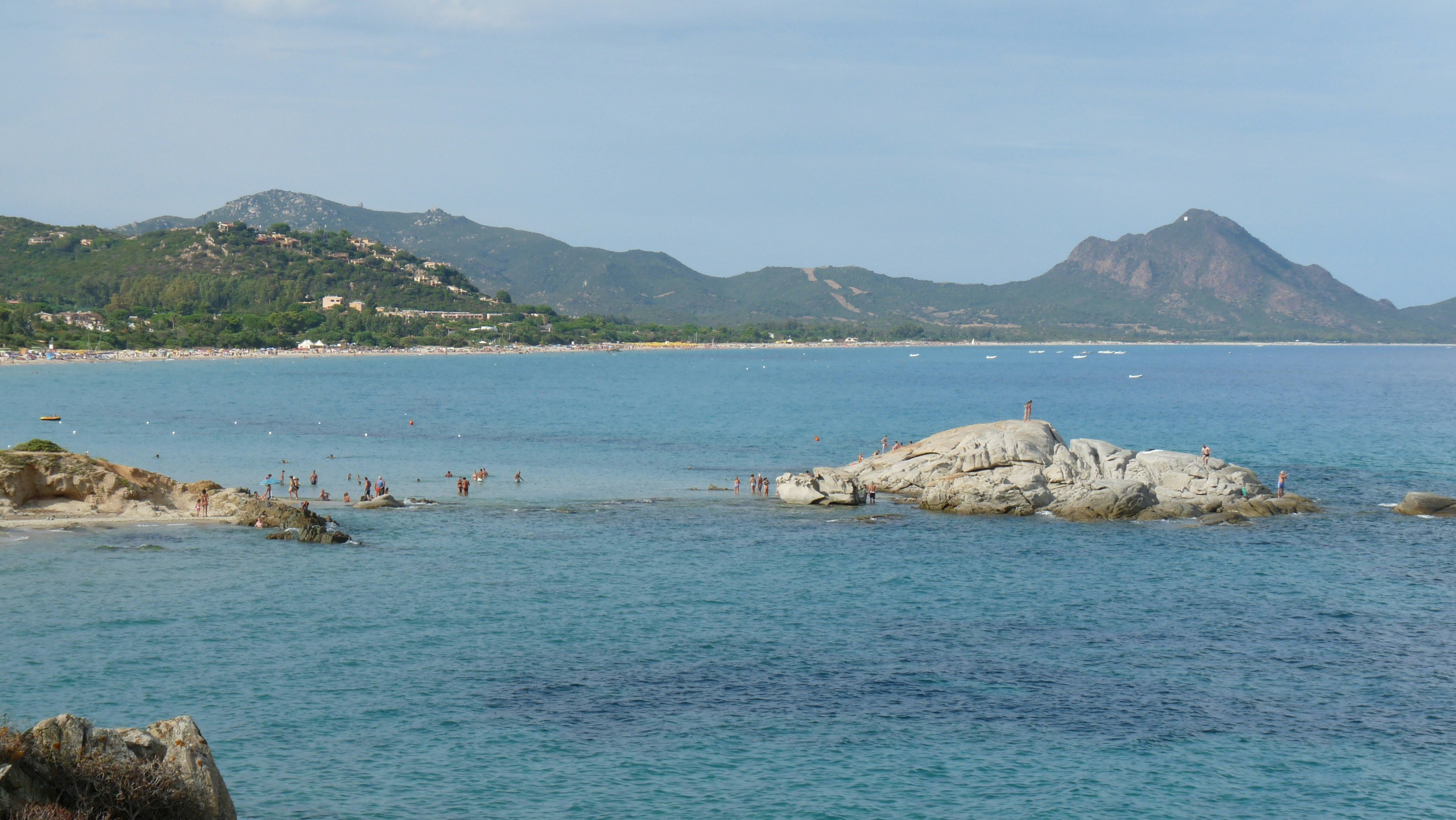 Blue ocean with rocks, a sandy beach, and distant mountains.