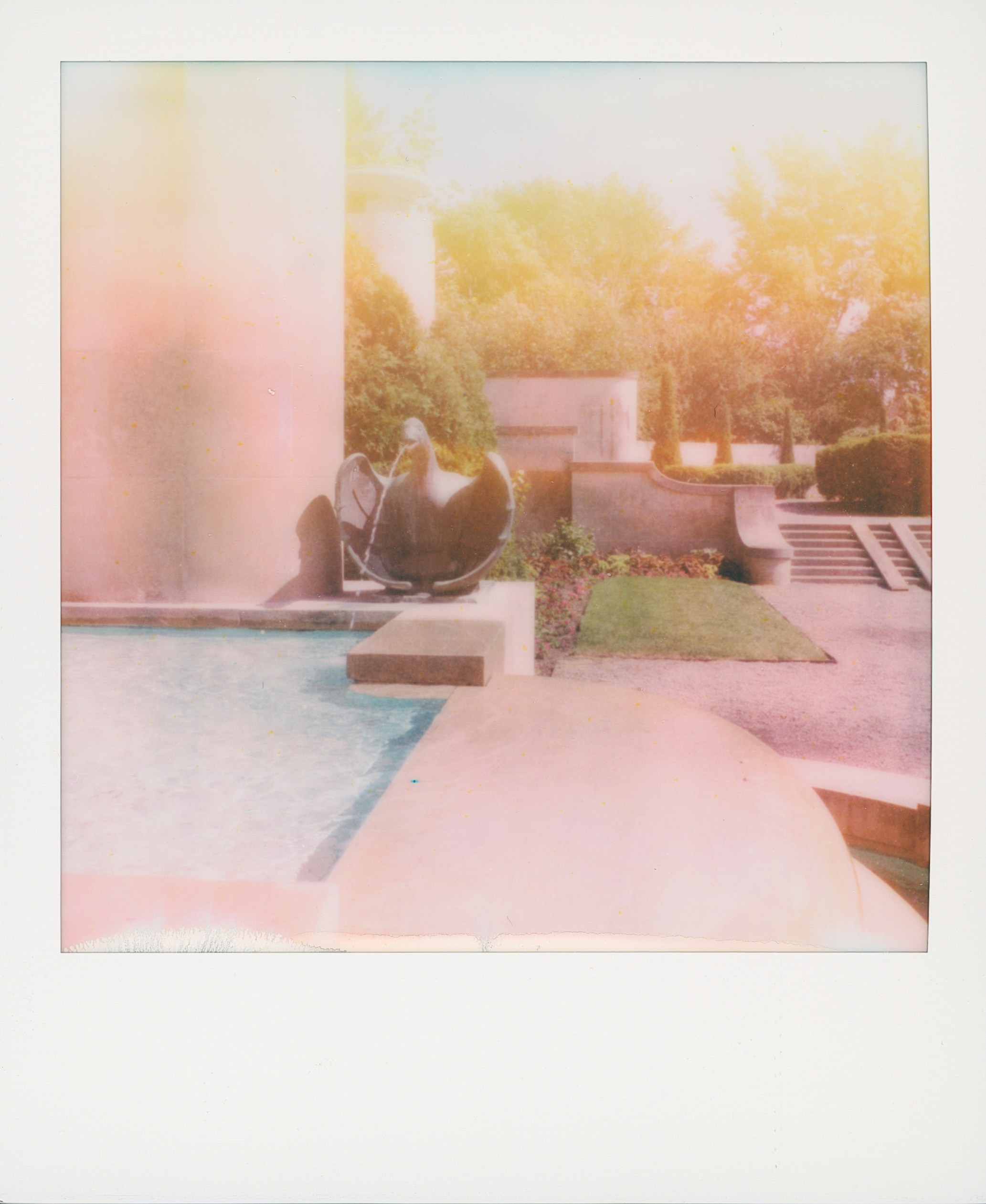 Faded outdoor scene with pool, sculpture, grass, and architectural elements.