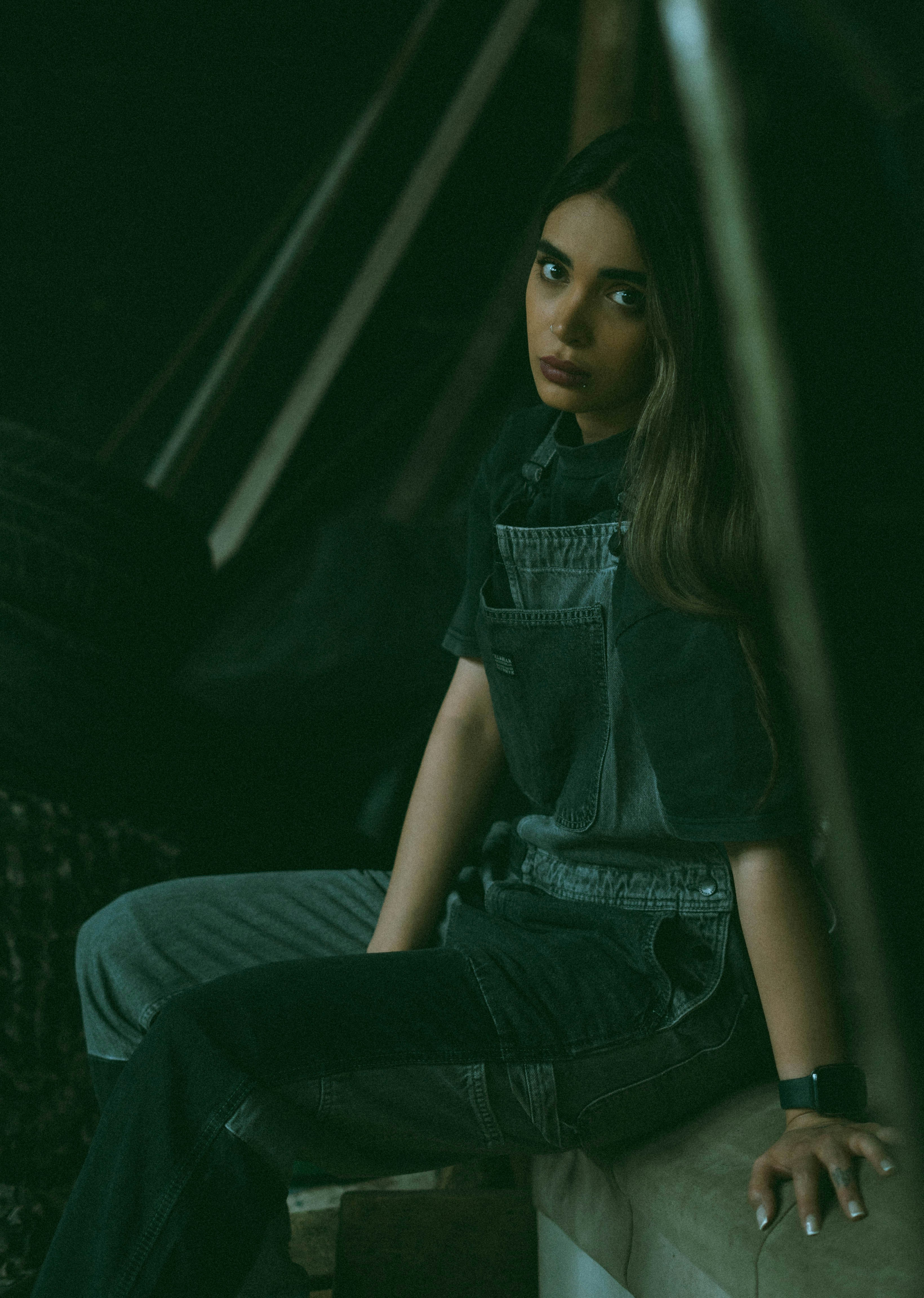 Mulher de macacão jeans sentada em um interior escuro e melancólico.
