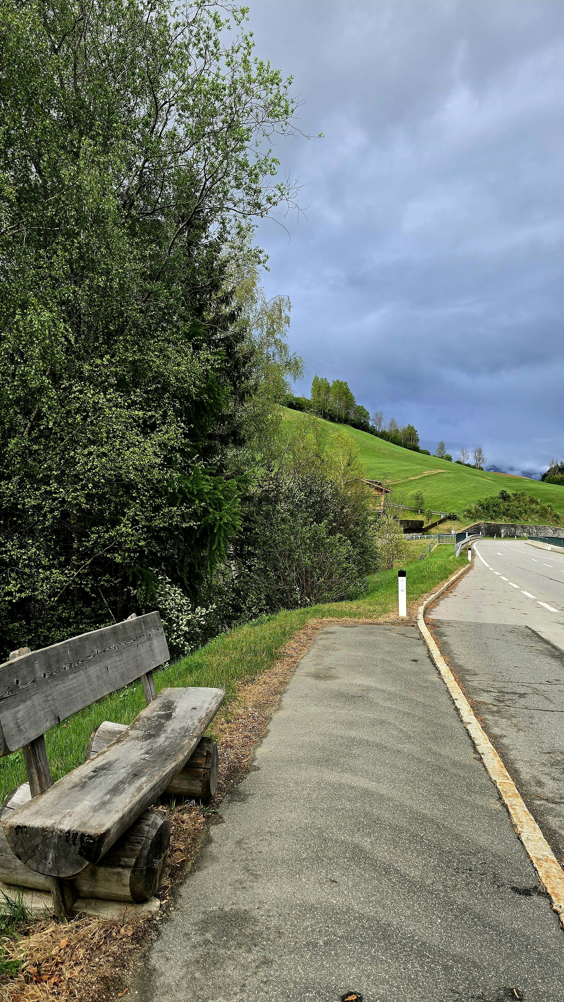 Empty wooden bench next to a road and green hillside