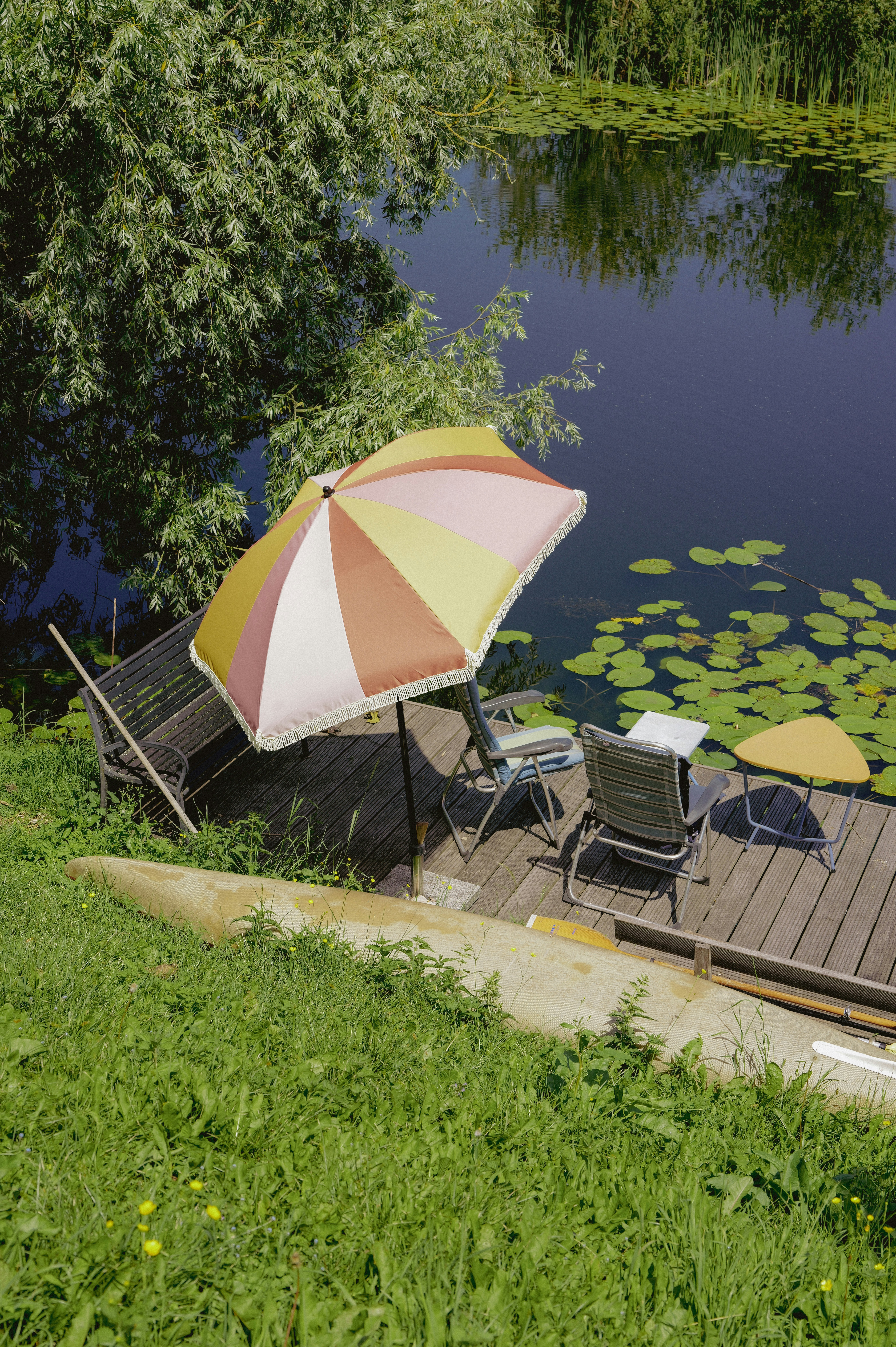 Relaxing dock with umbrella, chairs, and lily pads on water.