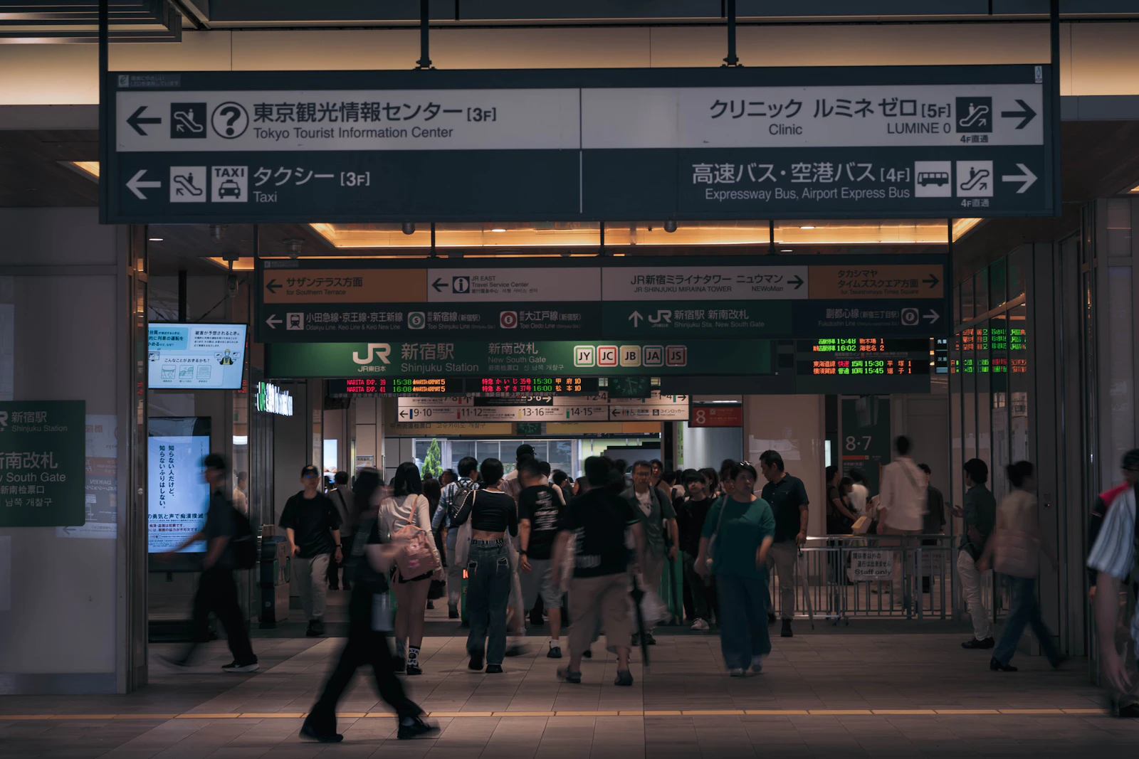 Photograph of people pass below overhead direction signs in a large rail-station concourse.