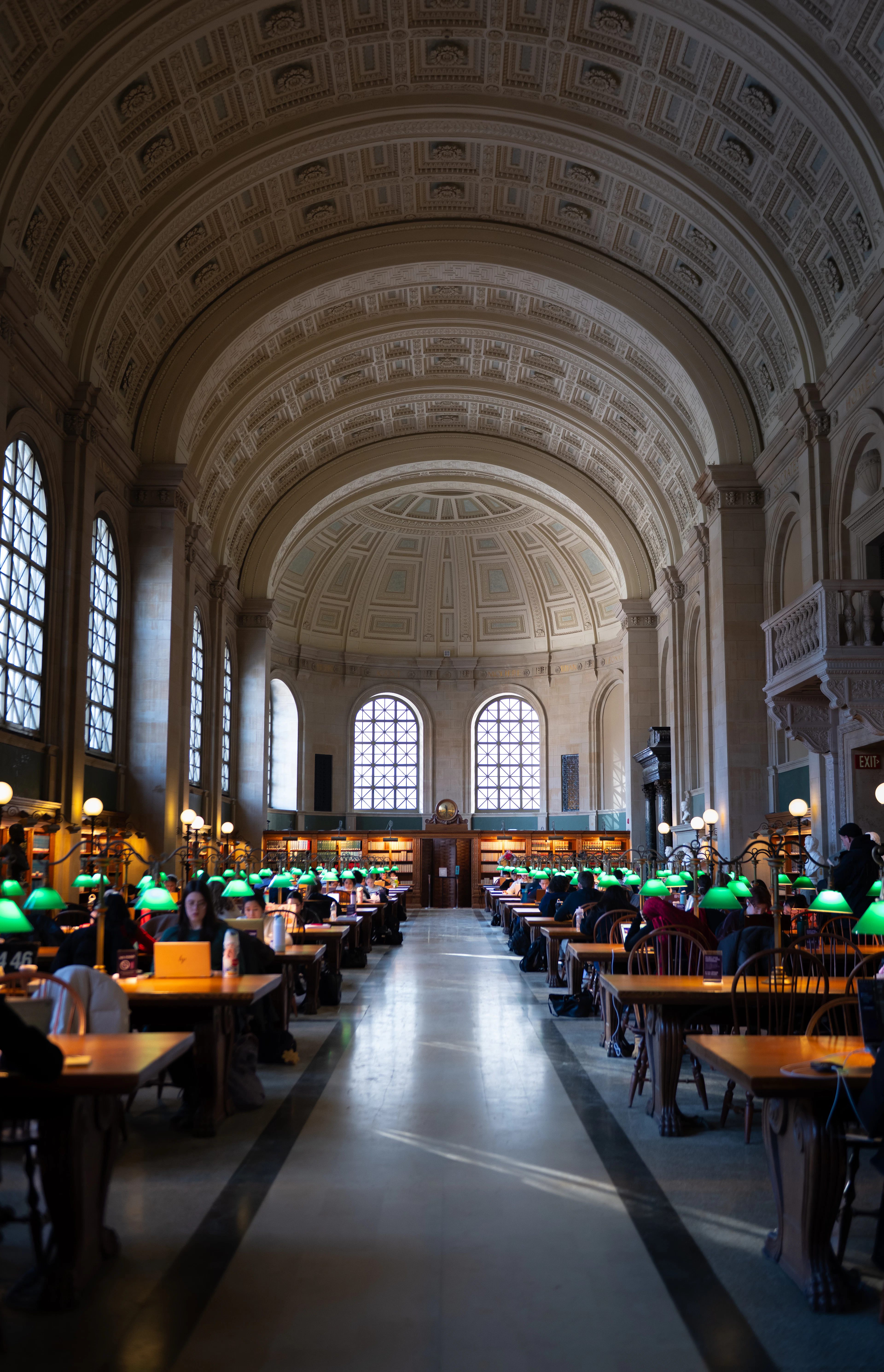 The barrel-vaulted reading room of the Boston Public Library, its long oak tables lit by rows of green banker's lamps beneath tall arched windows and a coffered stone dome.