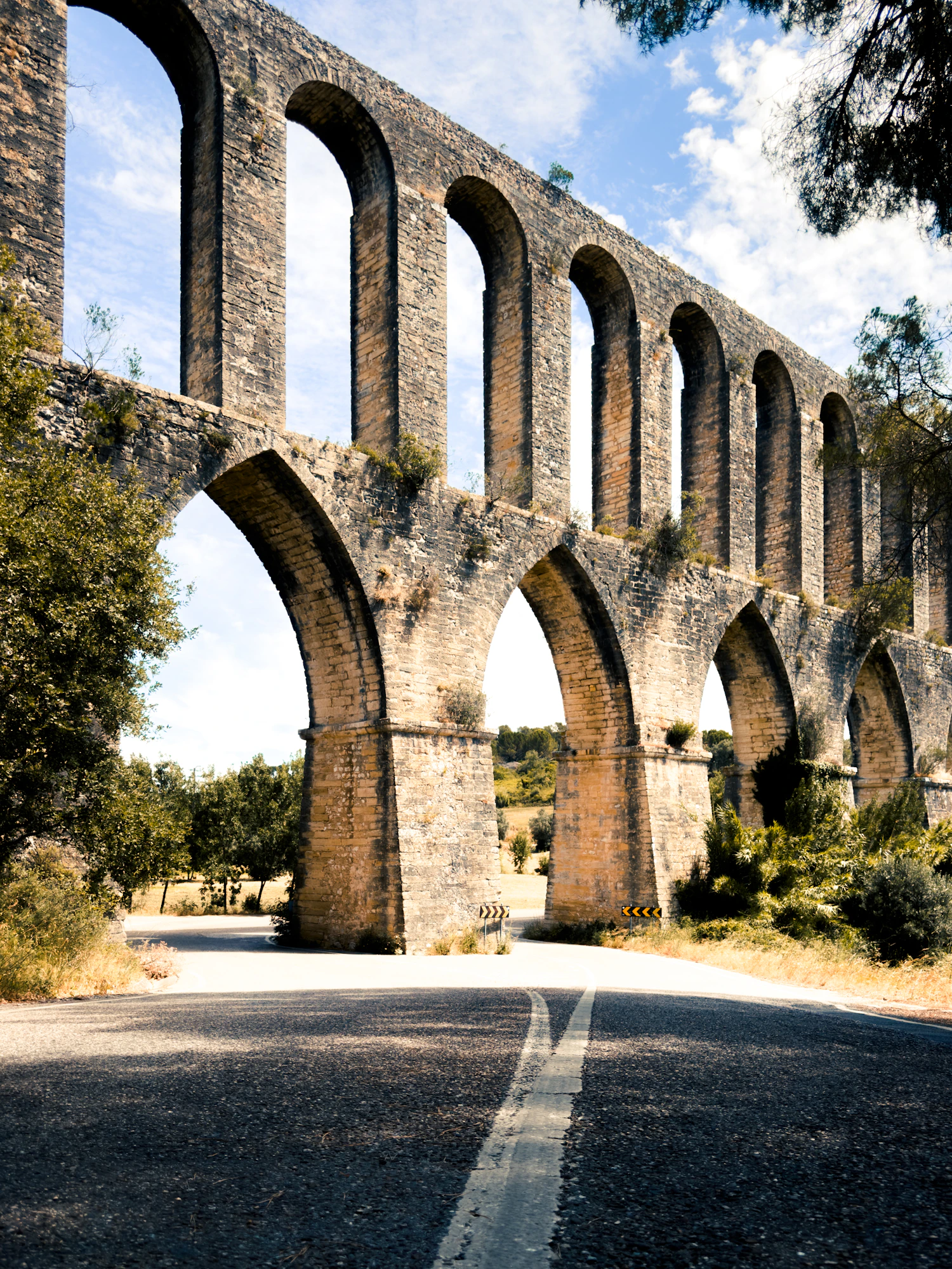 An ancient stone aqueduct arches over a road with trees.