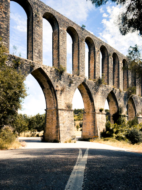 An ancient stone aqueduct arches over a road with trees.