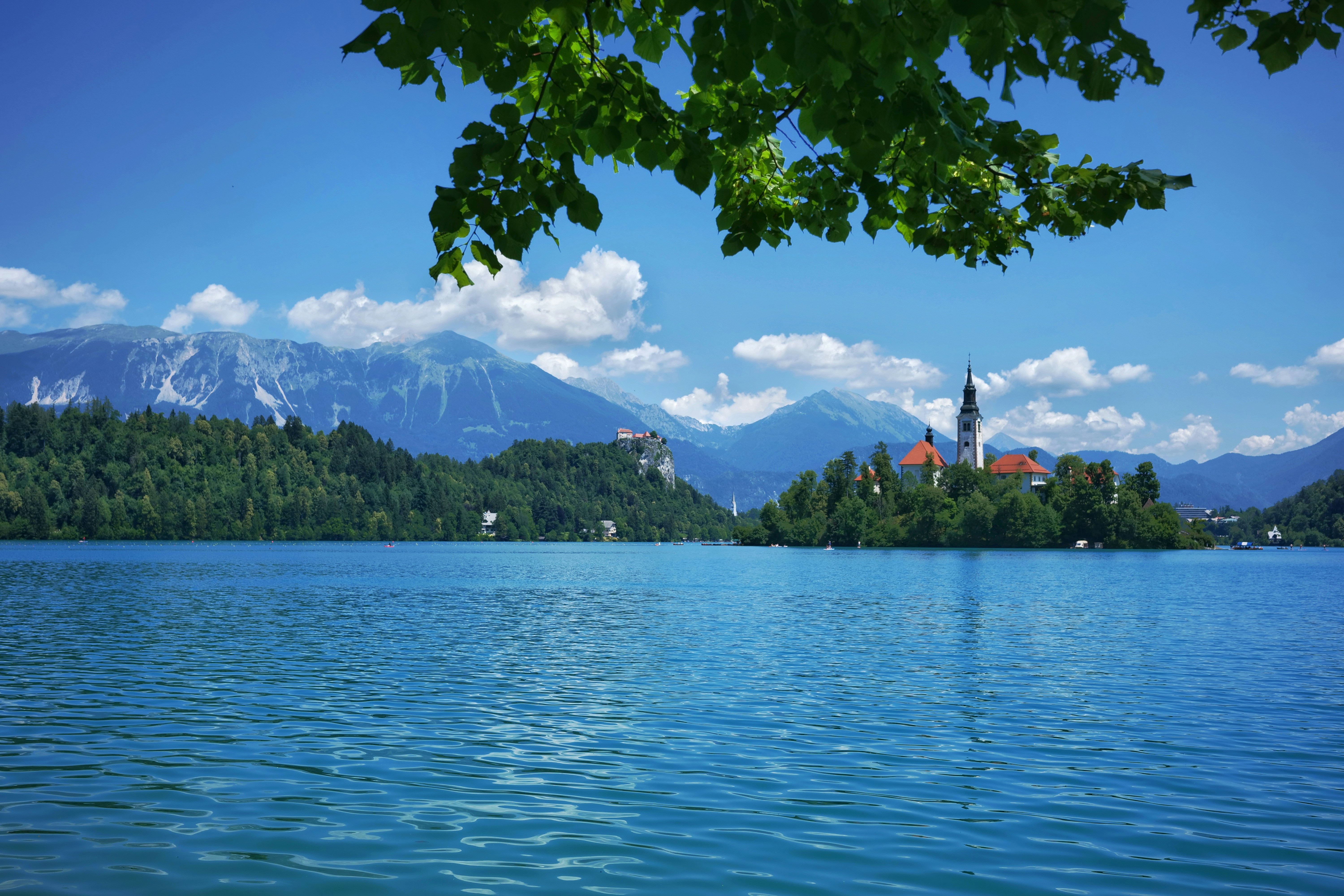 Blue lake, island church, castle, and mountains under clear sky.