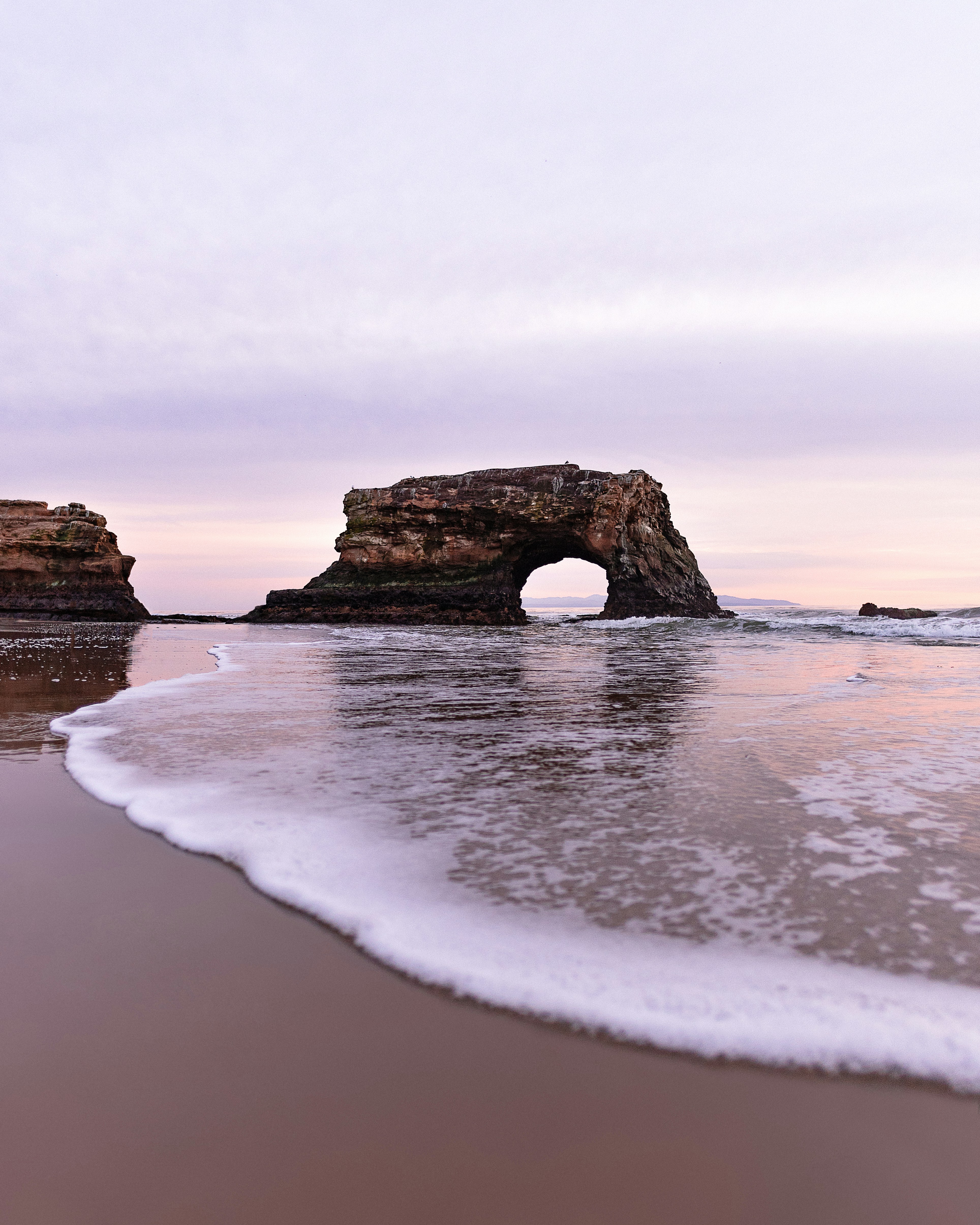 A natural rock arch stands in the ocean at sunset.