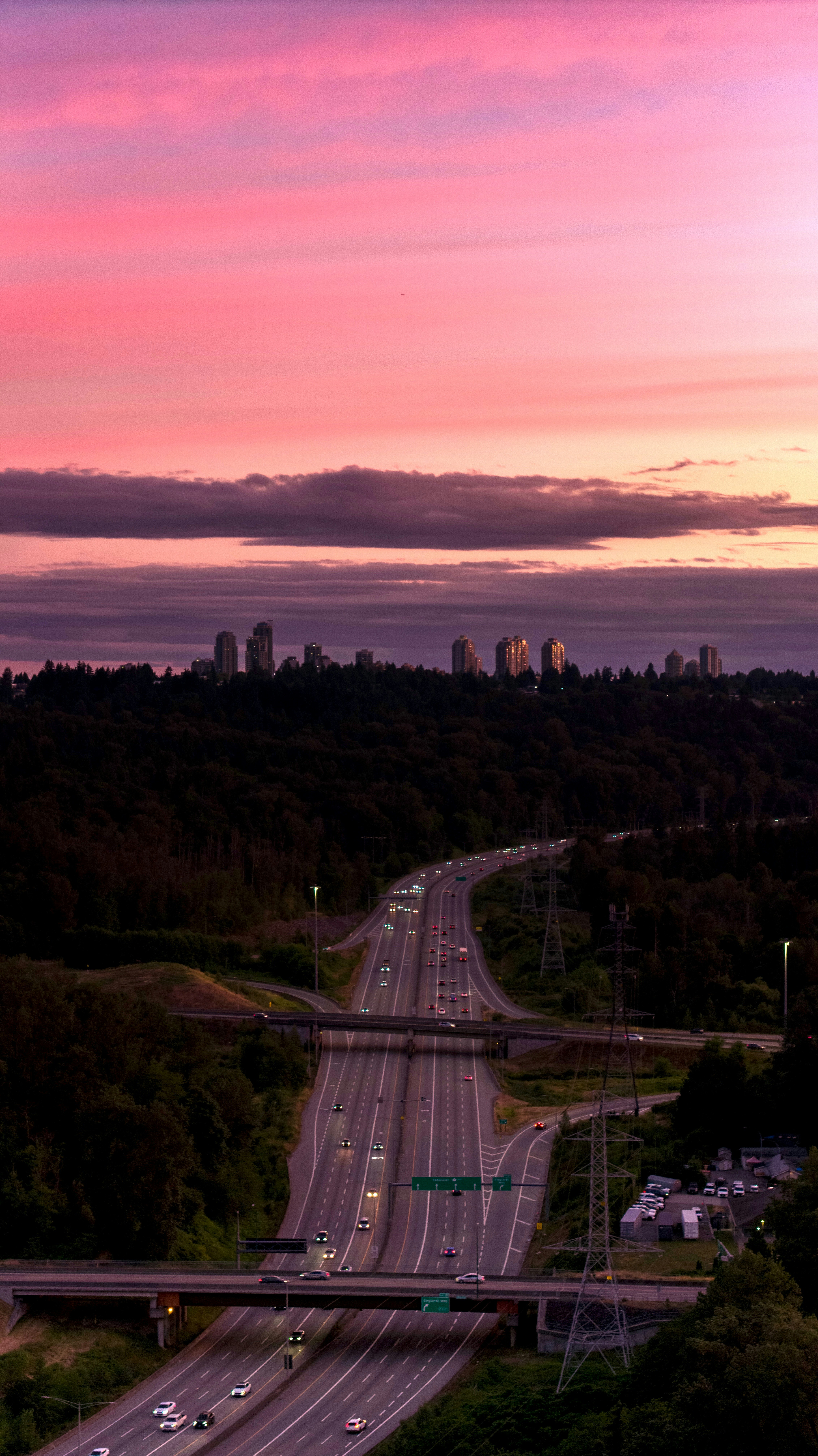 Vibrant pink sunset over highway, city skyline, and dark forest.