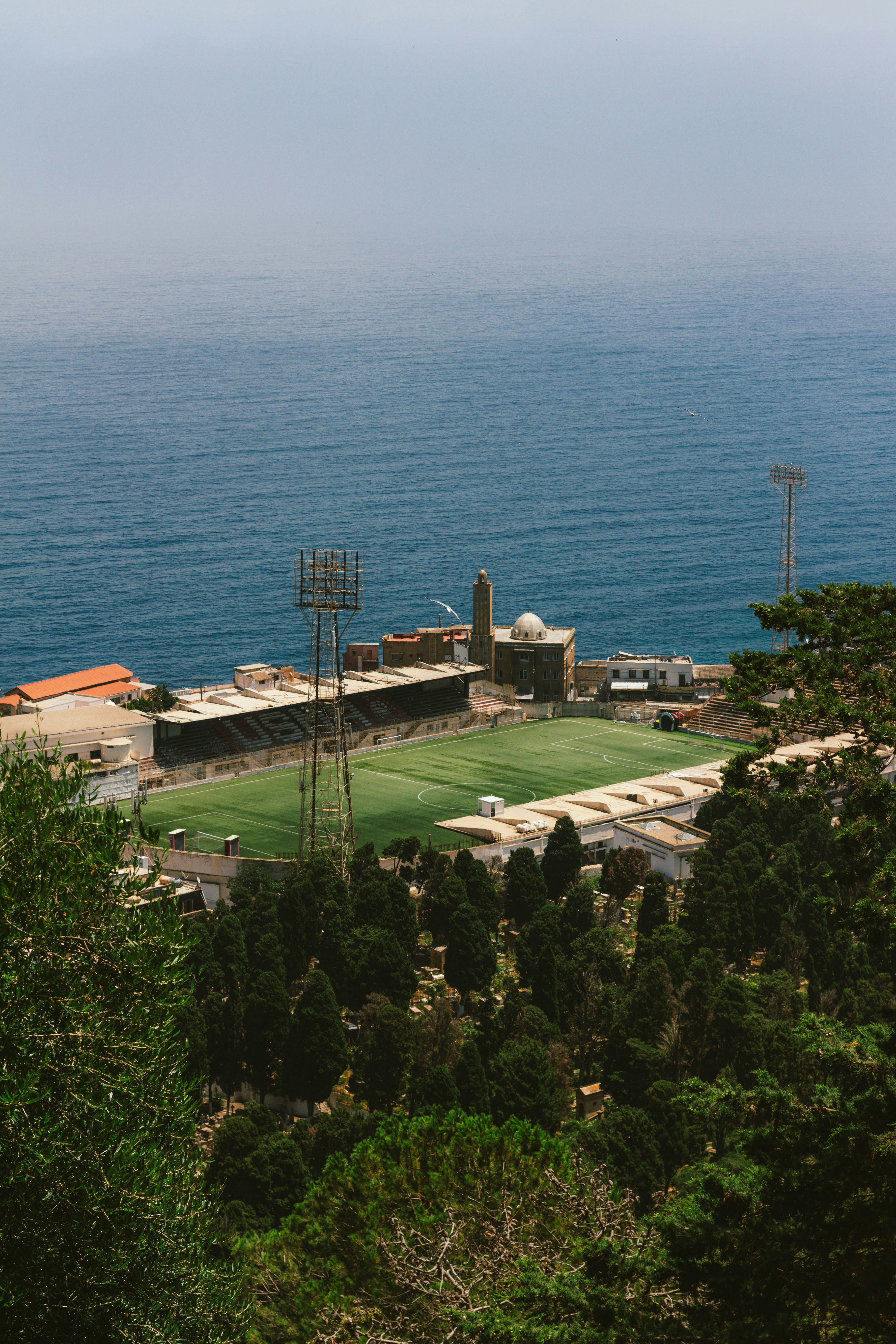 Estadio de fútbol costero rodeado de árboles con vistas al mar azul.